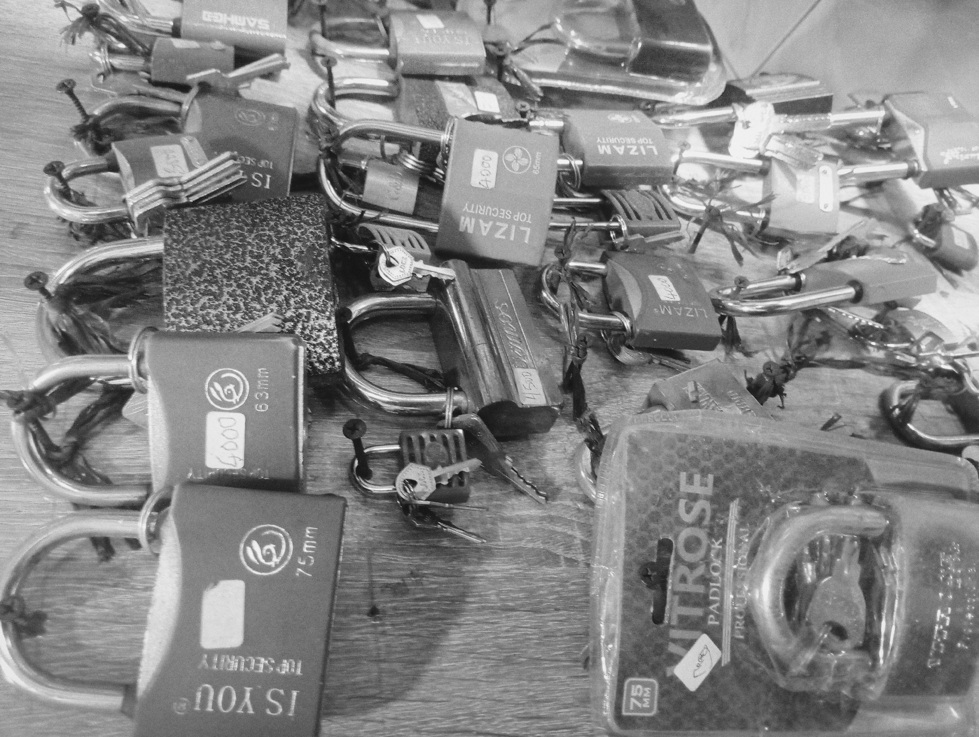 The image is a black and white photograph depicting a collection of padlocks spread across a wooden surface. Various brands are visible, including "IS YOU", "LIZAM", "SAMHED", and "KITROSE." The padlocks vary in size, some displaying measurements such as "63mm" and "75mm." Some padlocks are still in their original packaging, while others are loose, some with keys present. The scene appears to be indoors, with the wooden surface suggesting a table or countertop.