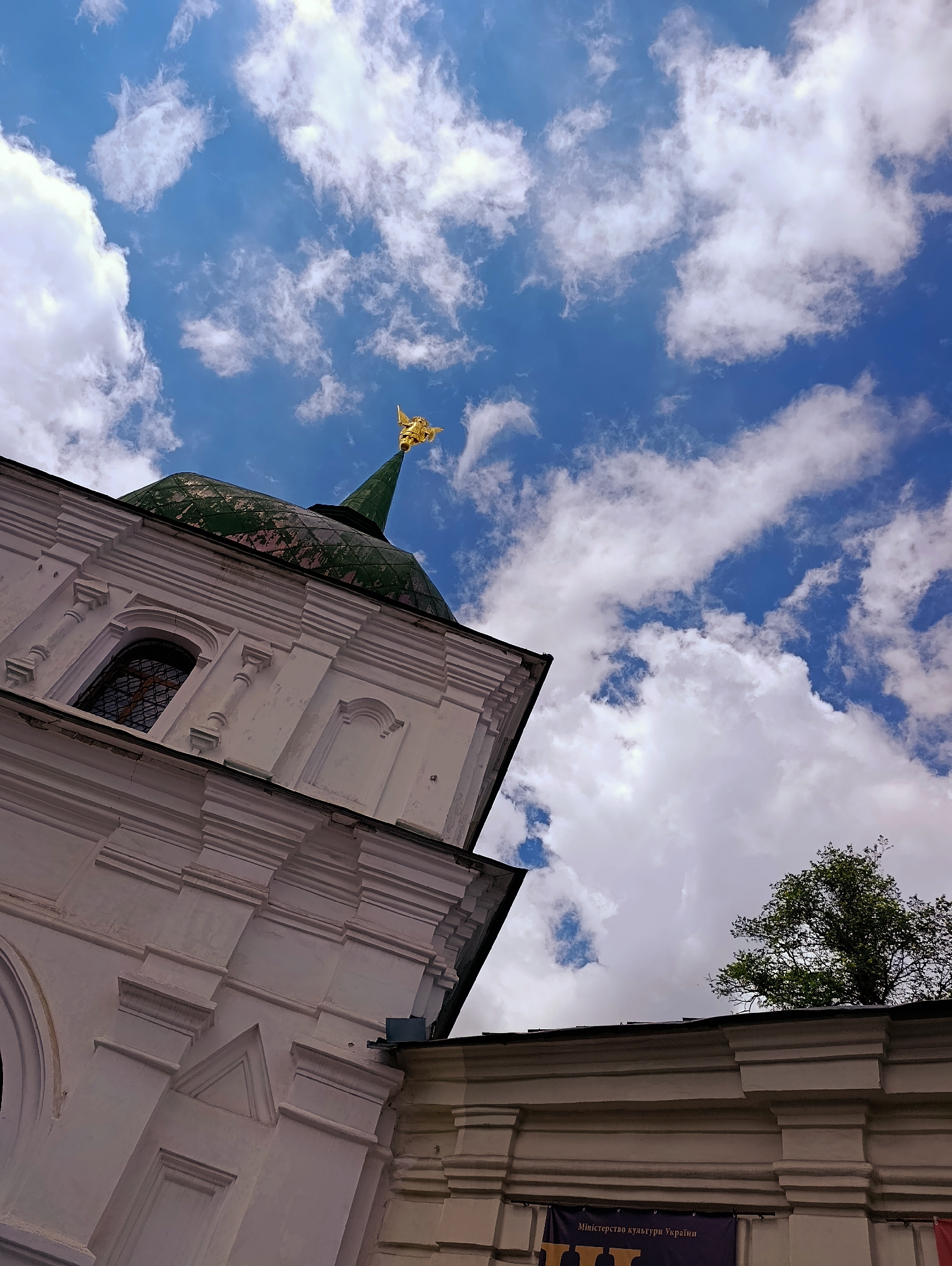 This is a low-angle, outdoor, eye-level shot of a white building with architectural details and a green domed roof topped with a golden cross. The building is part of a historic complex in Kyiv, Ukraine. The sky is blue with white, puffy clouds, and the sun appears to be shining. The building's facade is white with visible layers of molding and decorative elements. A dark-paned window is visible on the side of the building. A tree with green leaves is partially visible in the upper right corner of the image, behind the building. A banner with Ukrainian text is visible at the bottom of the image.