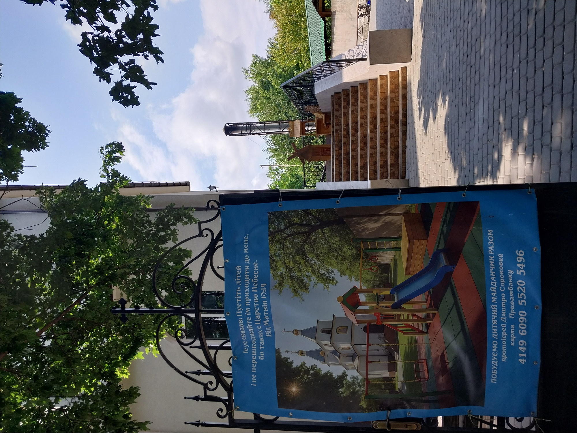 The image shows a large blue banner hanging on a black metal fence in Doslidne, Ukraine. The banner features a picture of a church and a children's playground. The text on the banner is in Ukrainian and appears to be a religious quote followed by information about building a playground.
The banner is the primary subject, occupying the left half of the frame. It is attached to the fence with visible grommets and ties. The fence itself is ornate with scrollwork. Above the banner and fence, the branches of a large tree with green leaves are visible against a blue sky with scattered clouds.
To the right of the fence and banner, a set of outdoor stairs with patterned tile risers leads up to an elevated area. A black metal railing runs alongside the stairs. On the landing at the top of the stairs, a wooden decorative windmill and a metal chimney are visible. Beyond the stairs and railing, green trees and a building with a green roof can be seen. The ground in front of the fence is paved with grey interlocking stones. The overall impression is that of a religious or community space with a focus on children's activities.