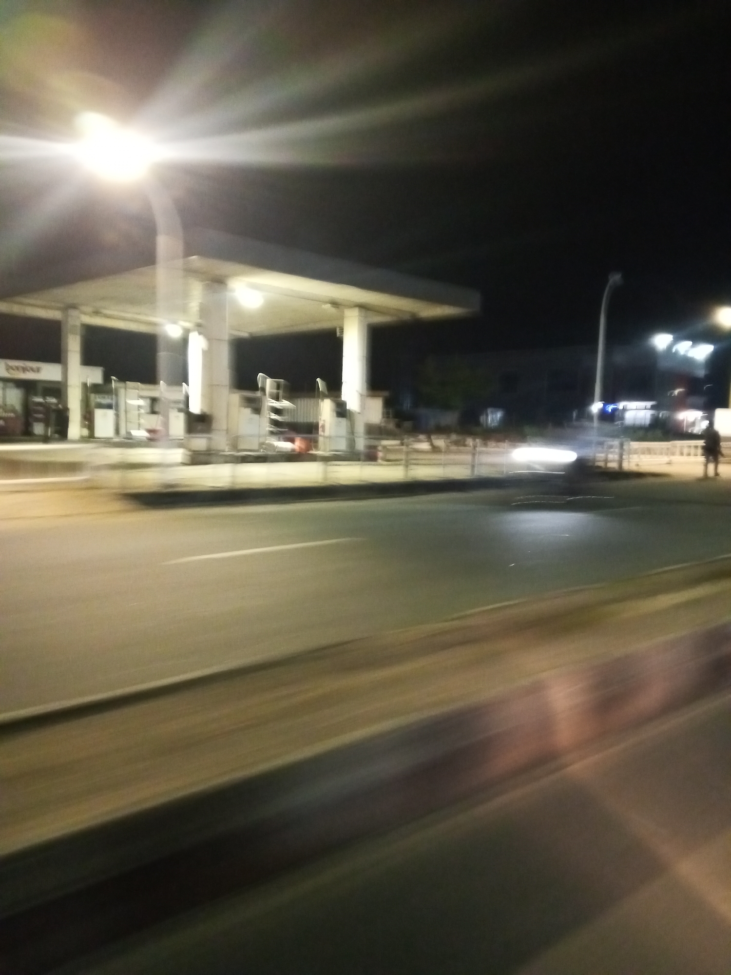 The image depicts a motion-blurred nighttime scene of a gas station in Lokoja, Nigeria. A brightly lit gas station canopy with overhead lights is visible. Multiple fuel pumps are present under the canopy. A street runs in front of the gas station, with lane markings visible. In the background, there are buildings and streetlights. A person is standing on the right side of the frame. The overall impression is of a typical urban night scene at a service station, captured with a moving camera.