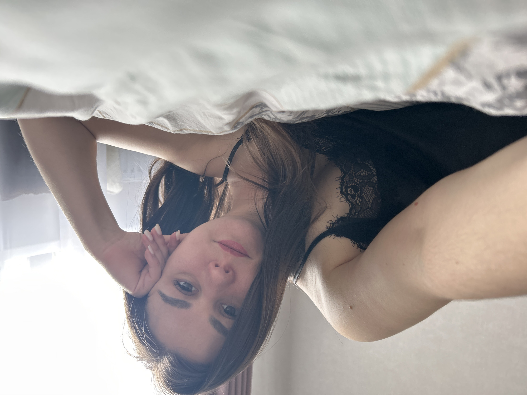 A woman with long brown hair is lying in bed. She is wearing a black lace-trimmed tank top. The image is taken from a low angle, looking up at her from under the covers. Her head is resting on her hand. The location is likely indoors. The scene appears to be in a private residence, possibly in the city of Chaikovsky, Russia.