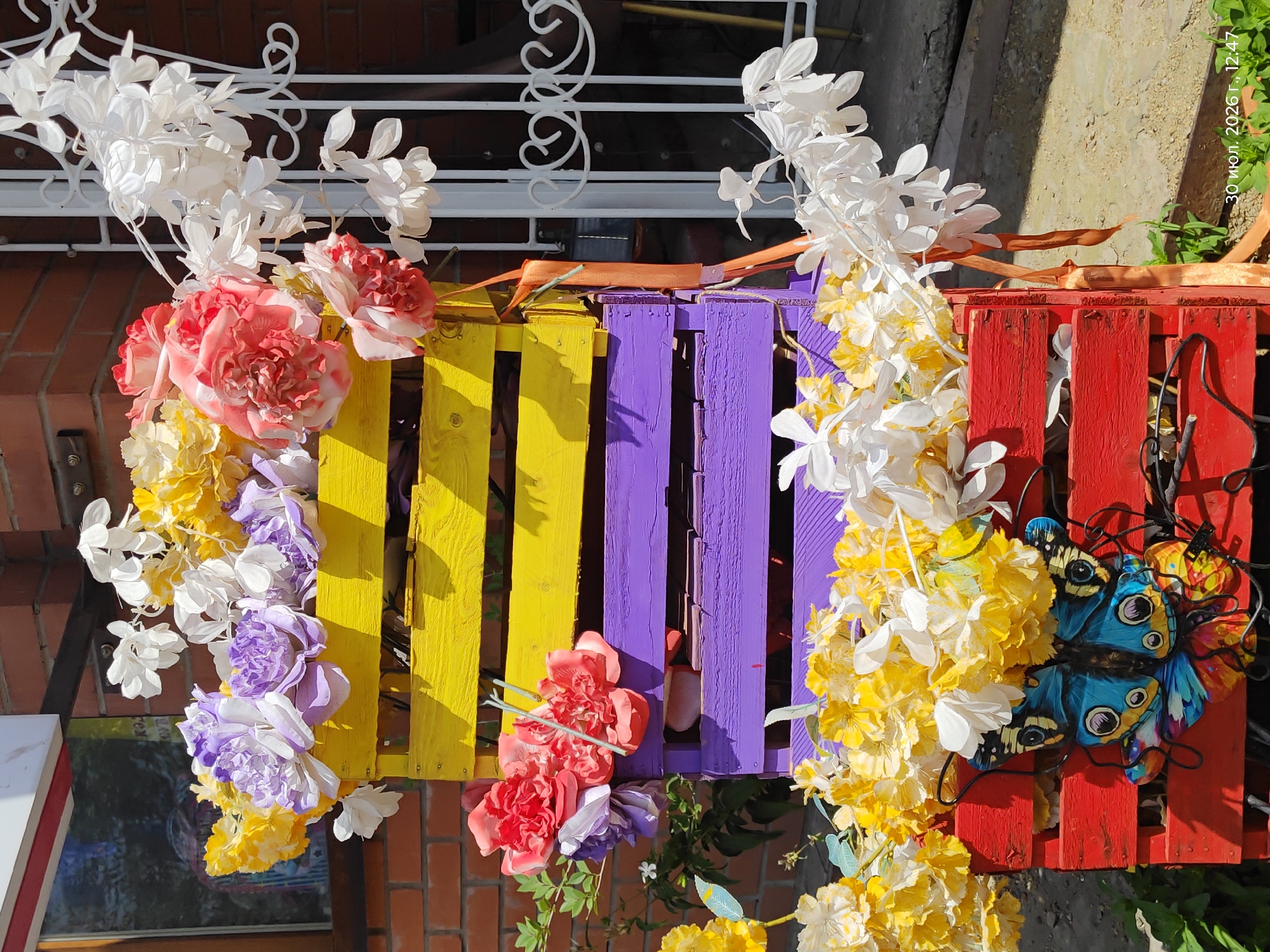 The image displays a decorative outdoor arrangement featuring stacked wooden crates painted in vibrant colors: red at the bottom, purple in the middle, and yellow at the top. These crates are filled with artificial flowers in various colors, including yellow, white, purple, and pink. A metal butterfly decoration is affixed to the front of the red crate. The arrangement is situated outdoors, with a brick wall and a white metal gate visible in the background. The ground appears to be a mixture of dirt and gravel. A timestamp in the lower right corner indicates "30 июл. 2026 г., 12:47". There are no people or animals present in the image. The scene is likely in Astana, Kazakhstan, given the Cyrillic text in the timestamp.