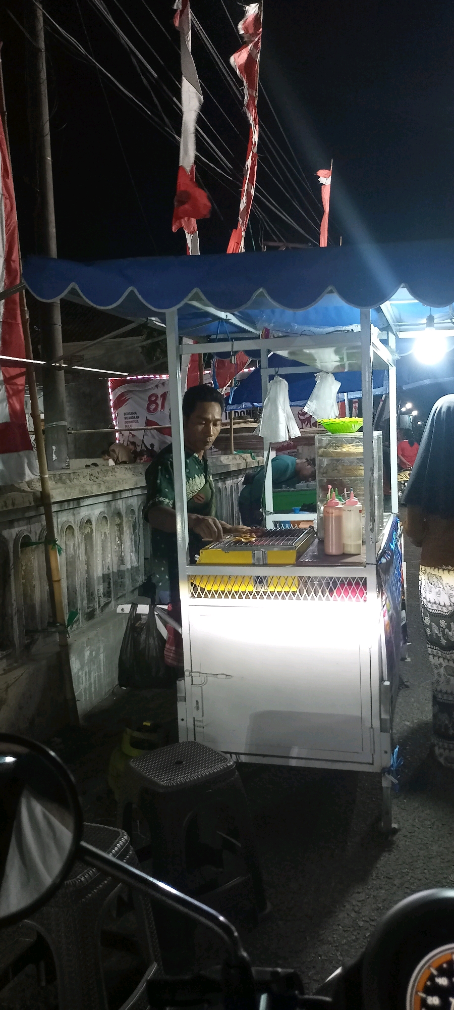 A nighttime street scene in Kalimati, Indonesia, captures a vendor operating a food cart. The cart is illuminated by a bright light, highlighting a grill where skewers of food are being cooked. Two bottles of sauce, one red and one white, sit on the counter of the cart. A man, presumably the vendor, is seen attending to the grill, wearing a green patterned shirt. In the background, several red and white Indonesian flags are visible, some tattered and blowing in the wind. The street appears to be paved. Other individuals are present in the background, though their details are obscured by the darkness and distance. Part of a motorcycle's side mirror is visible in the foreground, suggesting the photographer is on a motorcycle. The overall atmosphere is that of a bustling night market or roadside food stall.