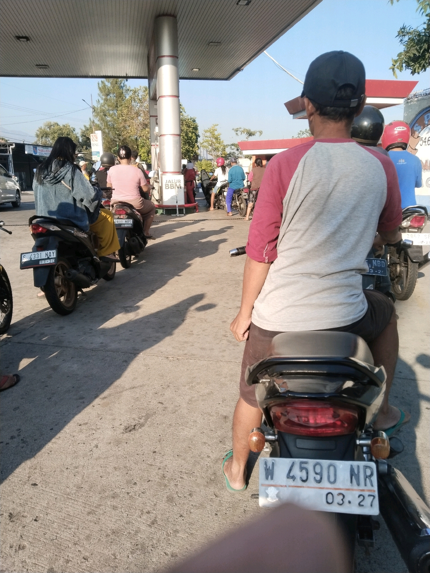 The image captures a scene at a gas station in Pandanarum, Indonesia, on a bright, sunny day. The foreground is dominated by the rear of a black motorcycle with a rider wearing a maroon and grey t-shirt and a black baseball cap, facing away from the camera. The motorcycle's license plate reads "W 4590 NR" and "03.27". Behind this motorcycle, another motorcycle is visible, also with a rider. In the mid-ground, several other motorcycles and riders are lined up, some waiting to refuel. One motorcycle, in the left mid-ground, has a license plate "S 4331 NAY" and "11.30".
The gas station features a prominent canopy supported by a silver pillar with red accents, and a white sign that says "JALUR BBM". Several individuals are seen on motorcycles, some wearing helmets and casual clothing, including a woman in a denim jacket and yellow pants on the leftmost motorcycle and a person in a pink shirt on a motorcycle behind her. A silver car is parked in the background on the left. The ground is paved with concrete, and shadows are cast across it by the canopy and the vehicles. The overall atmosphere suggests a typical day at a busy Indonesian gas station.