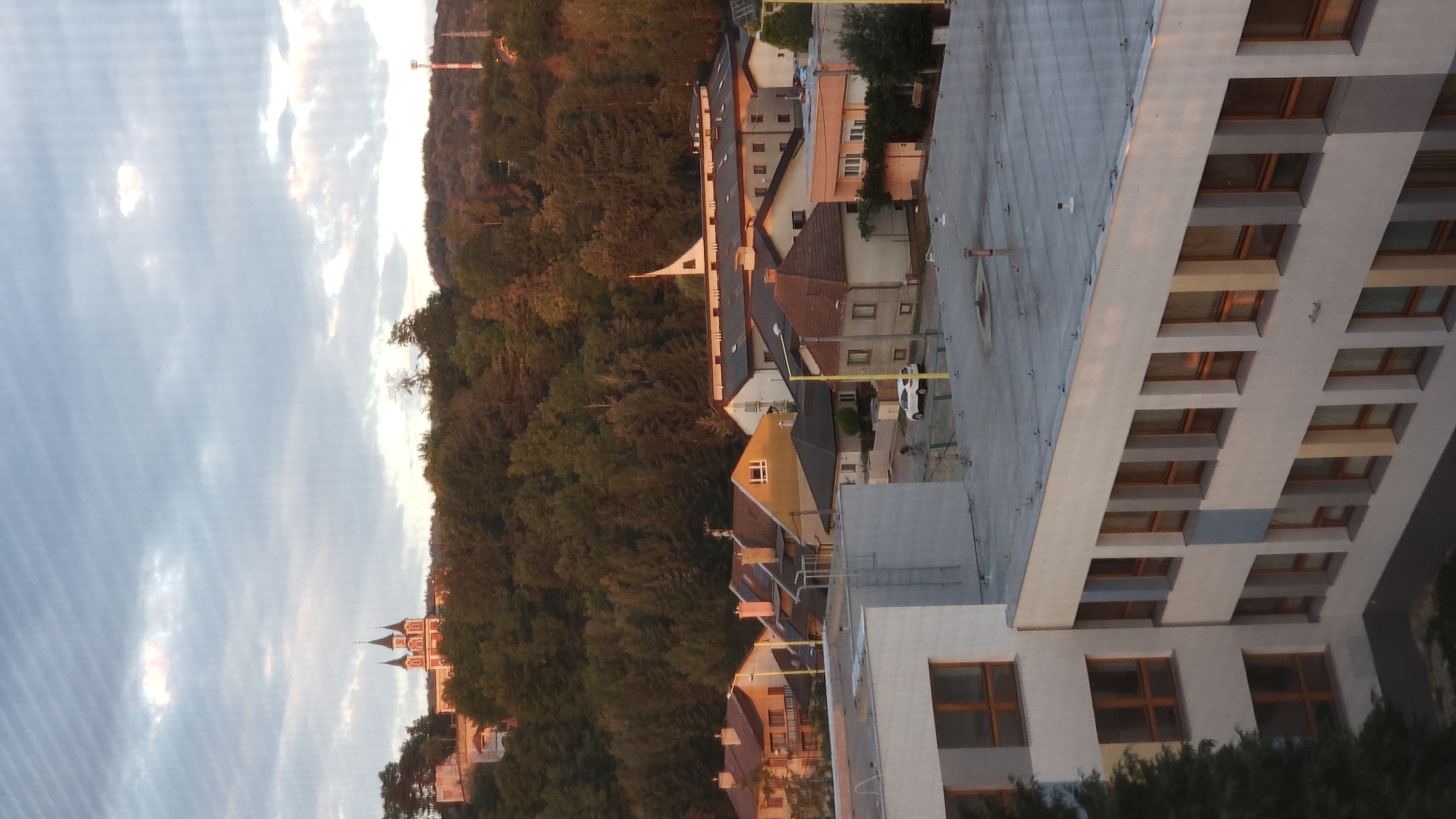 This is a vertical, eye-level shot of a town with residential buildings and a wooded hillside in the background. In the foreground, a multi-story building with a flat roof is partially visible on the left. This building has several rows of windows with dark frames. Further back, a cluster of houses with red-tiled roofs are seen, some with dormer windows. A white car is parked on the street between the houses. Beyond the houses, a densely wooded hill rises, topped by a building complex with a distinctive white spire. On the far left of the hill, a building with multiple towers is visible, possibly a church or historical monument. A tall telecommunications tower with red and white markings stands on the right side of the hill. The sky above is partly cloudy with a hint of sunset light illuminating the clouds. The scene suggests an urban environment adjacent to a natural landscape, likely in a town or city with hilly terrain.