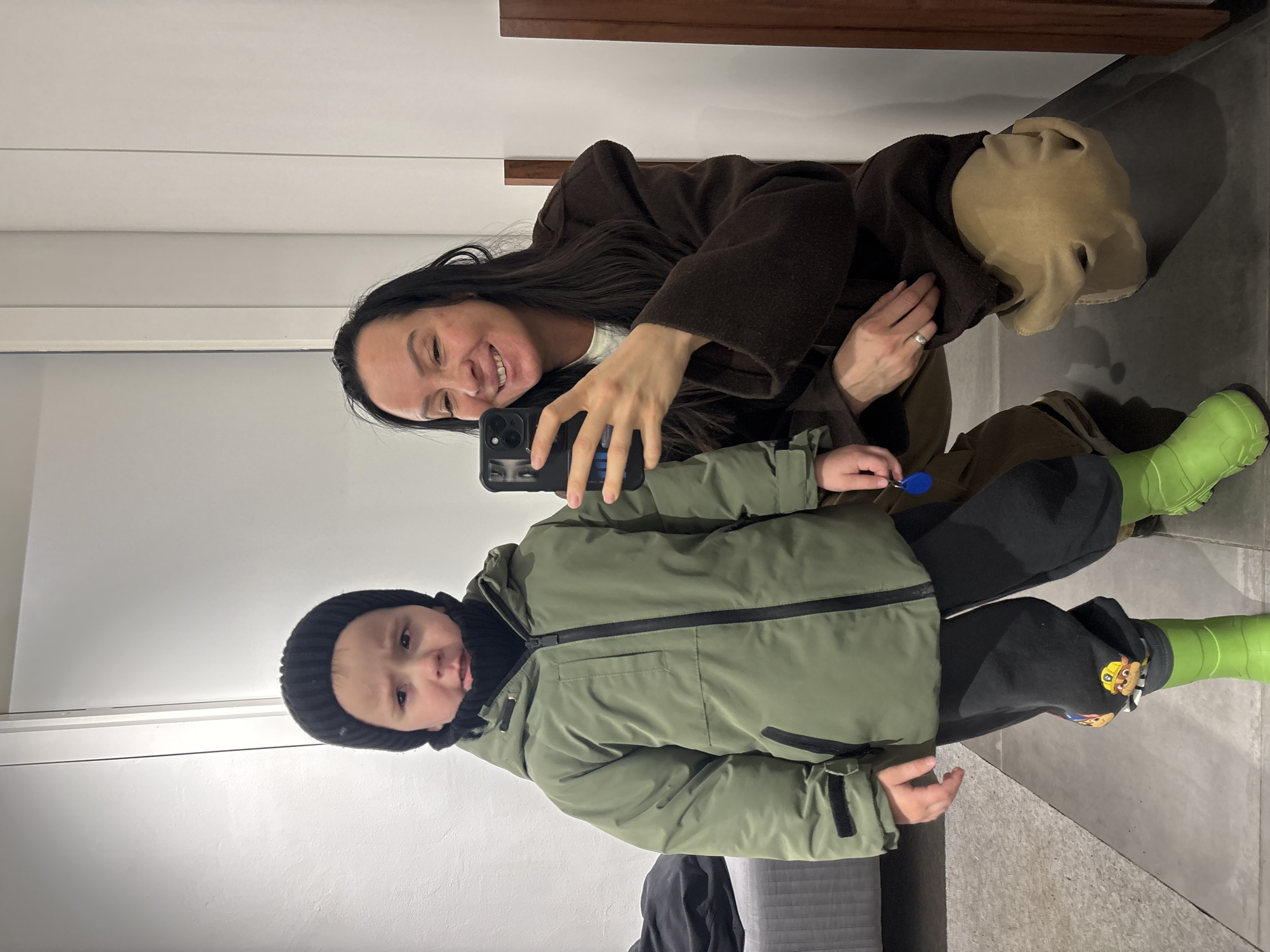 The image shows a selfie taken by a woman with long dark hair, wearing a brown coat. She is smiling and holding a black smartphone. In front of her is a young child wearing a black beanie, a green winter jacket, dark pants, and bright green rubber boots. The child appears to be looking at the camera. The location is Chaikovsky, Russia. The background appears to be an indoor space, possibly a store or fitting room with light-colored walls. There is a wooden shelf or fixture visible in the background. The lighting seems to be artificial. The child's clothing suggests it may be cold or wet outside. The child's pants have a cartoon character on them.