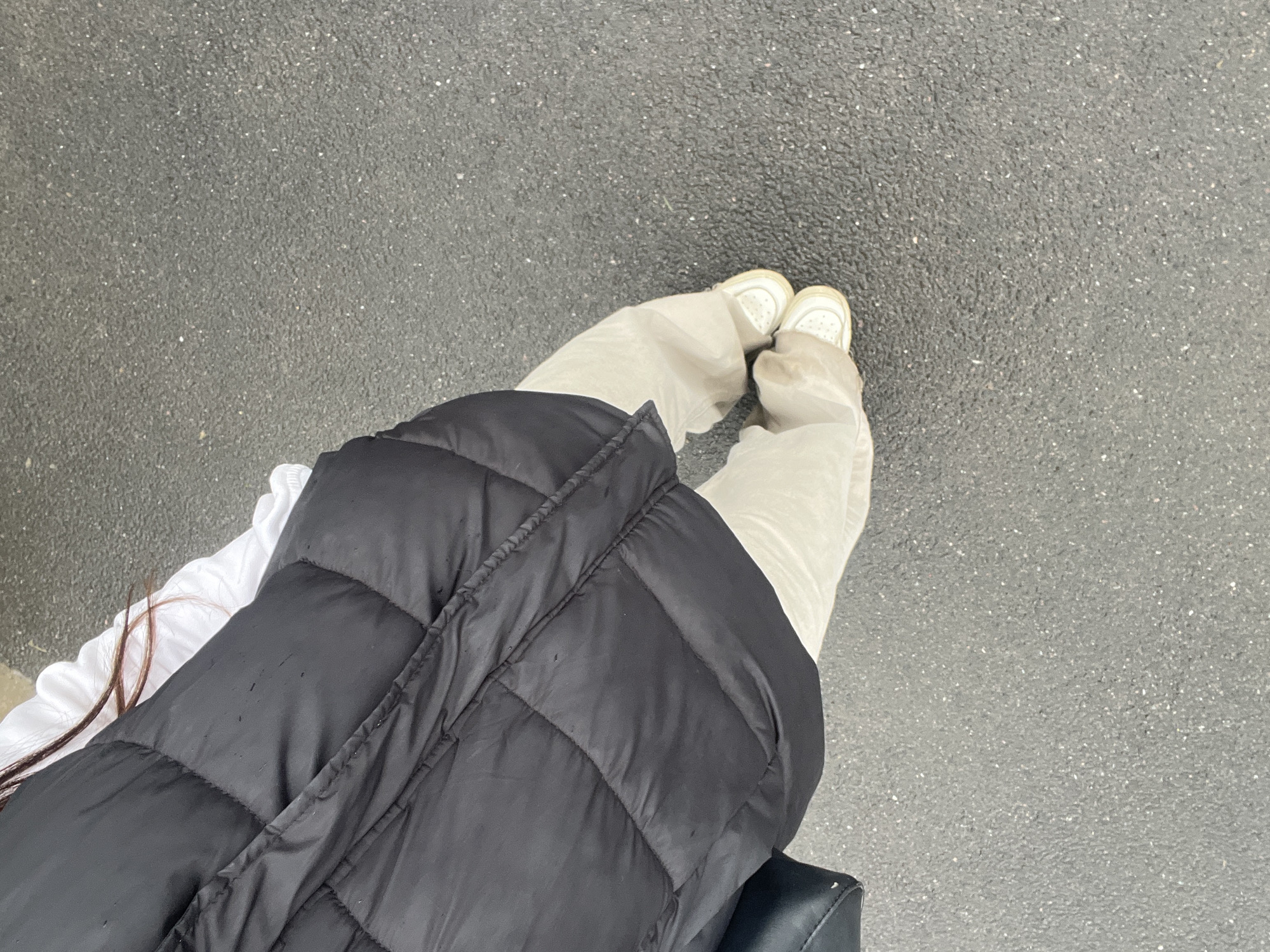 A person is sitting outdoors on a surface covered in asphalt. The individual is wearing white pants and white sneakers. A black puffer vest covers part of a white shirt. Two strands of brown hair are visible. The location is unknown.