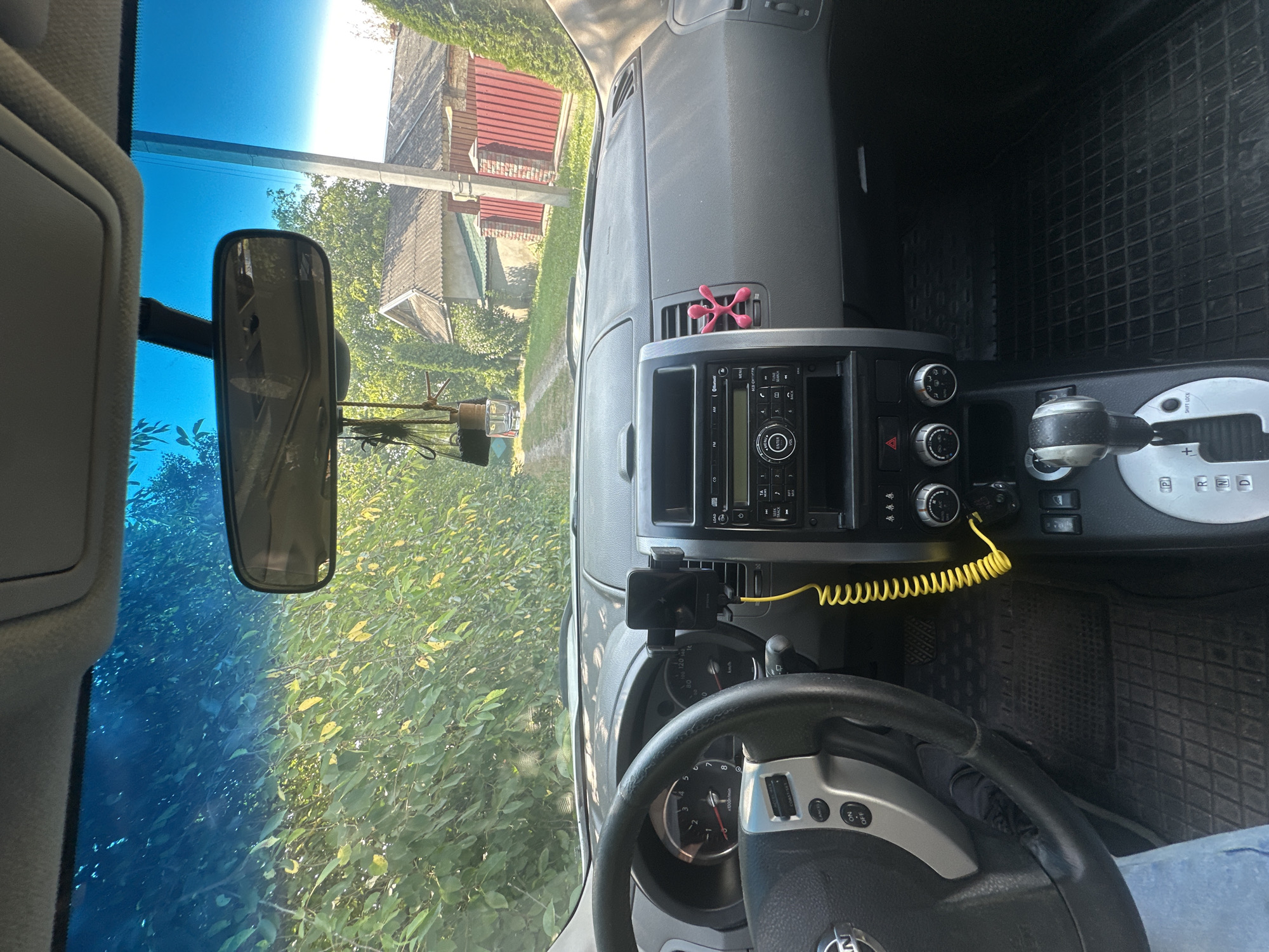 The image is taken from the driver's perspective inside a car, looking forward through the windshield. The car's dashboard is visible, including the steering wheel, instrument cluster, and center console with a car radio and gear shifter. A yellow coiled cable is attached to a phone mount on the dashboard. Outside the car, a dirt road leads past green foliage and houses. A red fence and a utility pole are also visible. The context provided suggests the image was taken in Shatava, Ukraine.