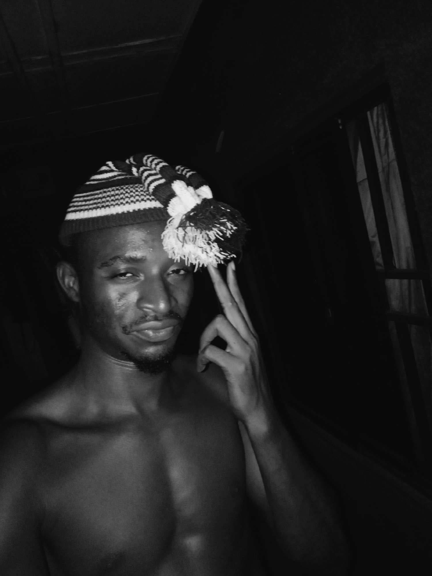 This black and white portrait captures a shirtless man, appearing to be of African descent, posing for the camera indoors in Onitsha, Nigeria. He is seen from the chest up, looking directly at the viewer with a slight smirk. On his head, he wears a striped knitted hat, featuring alternating patterns of thin and thick stripes in dark and light tones, and a fluffy pom-pom on the side.
His right hand is raised, with the index and middle fingers extended, making a "V" sign or peace gesture, and lightly touching the pom-pom of his hat. He has a trimmed beard and goatee, and his skin exhibits a slight sheen. The background is very dark, suggesting dim lighting, possibly nighttime. On the right side of the frame, faint details of what appears to be a window or door frame with vertical elements, resembling blinds or curtains, are visible. The image conveys a confident and direct demeanor. No visible text is present.