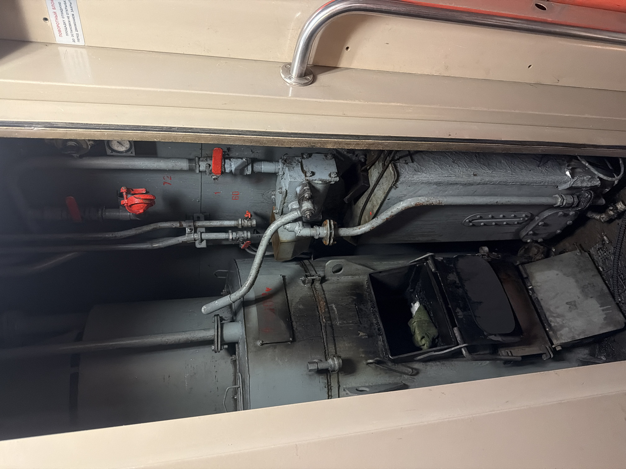 The image depicts a view inside a compartment that appears to be part of a boiler room or engine room, possibly within a train car or a stationary facility in Balashov, Russia. The compartment contains industrial equipment, including a large, cylindrical, grey metal tank on the left, likely a boiler or a water tank. In front of it is a smaller, dark-colored furnace or firebox with its doors open, revealing some debris inside, including a green cloth item.
To the right of the boiler, there are numerous pipes, valves, and gauges. One prominent red valve is visible, and another red valve is positioned above a gauge labeled "72." Further down, another red valve is near a gauge labeled "60." A rectangular, grey metal unit with bolts is mounted on the wall or equipment, with pipes connecting to it. Below this, another grey, ribbed rectangular tank is present, with pipes and fittings attached.
The compartment walls are painted a light beige color, and a section of a metallic door handle is visible on the far right. A warning sticker with Cyrillic text is affixed to the right wall, partially visible. The floor appears to be grimy and shows signs of ash or soot, consistent with a furnace or boiler operation. The overall impression is one of a functional, albeit somewhat aged and dirty, industrial space. The lighting is dim, contributing to the utilitarian atmosphere.