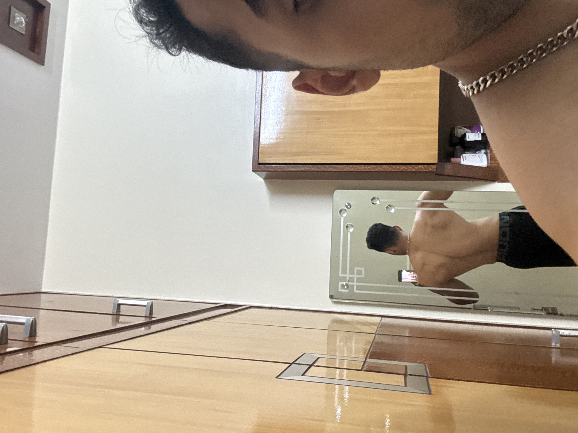 A person is taking a selfie in front of a mirror. The reflection in the mirror shows the person's back and a phone in their hand. The background includes a wall, cabinet, and a wooden floor with inlaid patterns. The location is inferred to be in Kolkata, India.