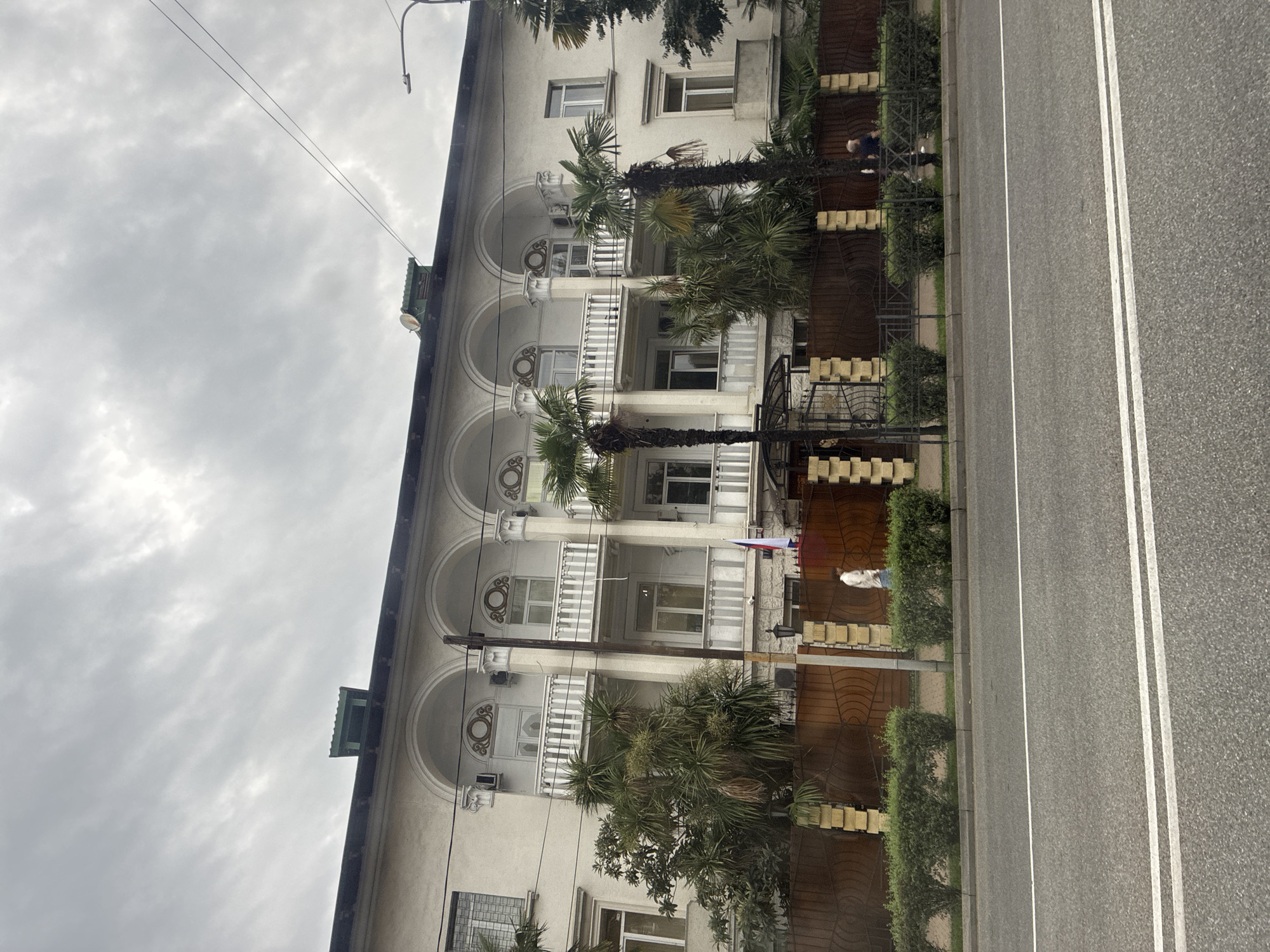 This is a medium-shot, eye-level photograph of a building in Sochi, Russia, on an overcast day. The building is light-colored, possibly stucco, with a dark green roof. It has a symmetrical facade with five arched openings on the upper floor, each with a balcony. Decorative circular motifs are visible above the windows within the arches.
In front of the building is a wooden fence with decorative metalwork and yellow pillar-like accents. A row of neatly trimmed green bushes runs along the fence line. Palm trees and other lush greenery are situated in front of the building and along the fence. A Russian flag is displayed on a flagpole to the right of the main entrance.
A person in a light-colored top and dark bottoms is walking along the sidewalk in front of the building. Further to the right, another person in dark clothing is visible near the fence. A street with white lane markings runs across the foreground of the image. A street lamp is visible on the right side of the frame. The sky is filled with dense, gray clouds. The architectural style suggests the building may be from the Soviet era, possibly a sanatorium or a government building, common in coastal resort towns like Sochi.