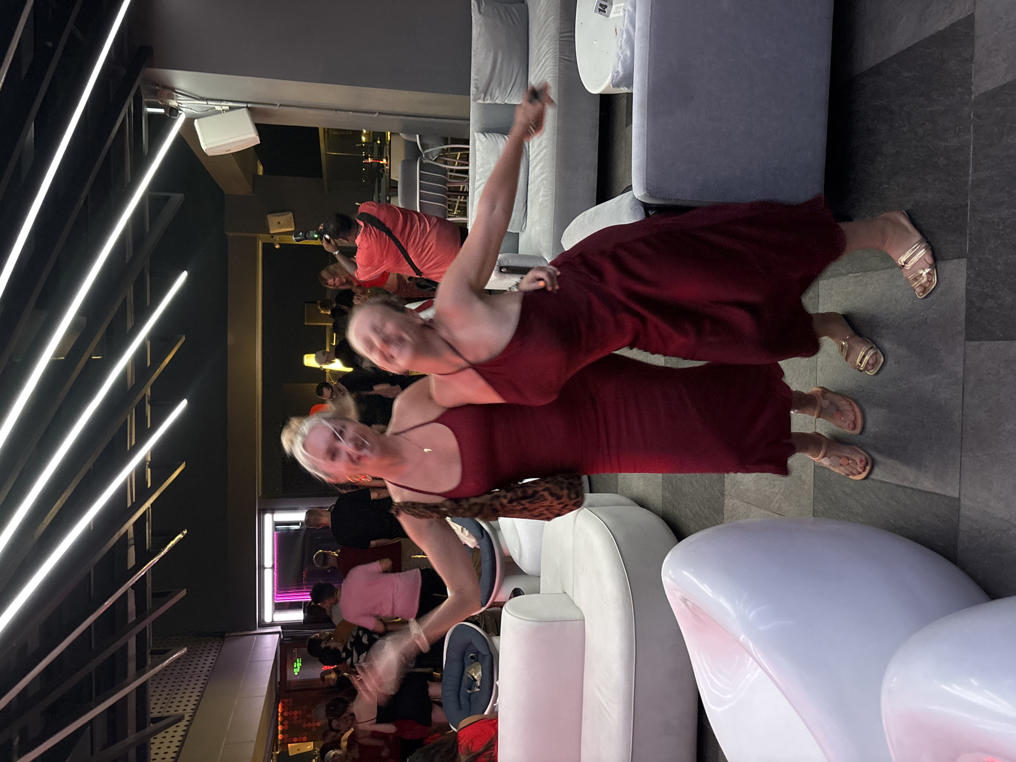 The image depicts a group of people inside a dimly lit establishment, likely a bar or club, with a photographer in the background taking a photo. Two women are prominently featured in the foreground, with one in a red dress appearing to be dancing or posing, while the other, also in a red dress, stands beside her. The woman in the red dress in the foreground has her arms outstretched and her face is blurred, suggesting movement. The woman behind her is smiling and looking towards the camera.
The scene is filled with bright, modern lighting from linear fixtures along the ceiling. The background shows lounge-style seating with plush grey cushions, and a white table. The floor is tiled with dark grey and black squares.
The time of day appears to be evening or night, given the artificial lighting and the indoor setting. There are no visible weather cues. The location is identified as Đà Nẵng, Vietnam. The overall mood seems to be lively and social, with people enjoying themselves.