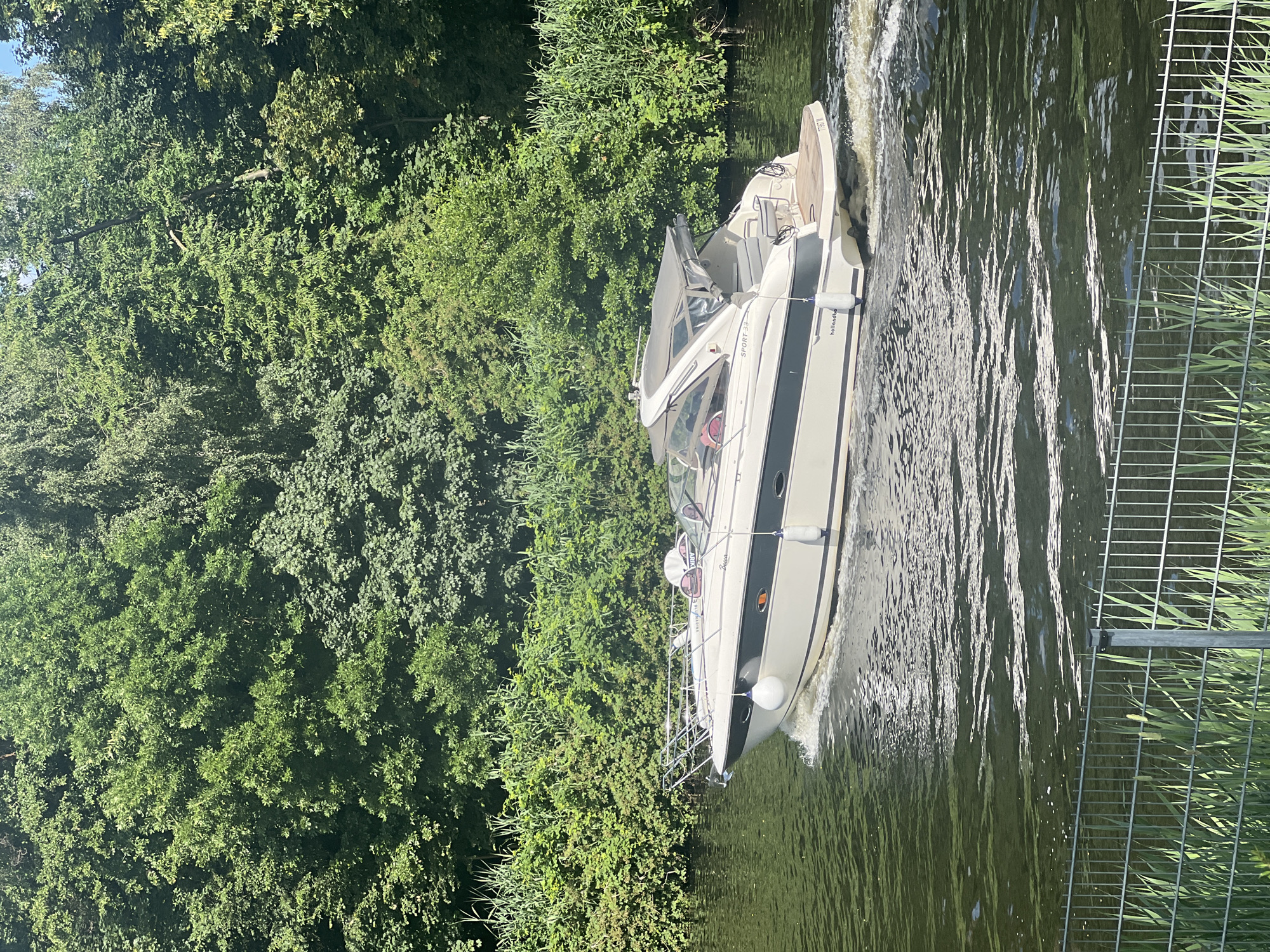 In Teltow, Germany, a white and gray motorboat travels on the Havel River. The boat has a covered cabin and a wooden deck at the stern, with white fenders attached to its sides. It creates a wake as it moves through the water. The riverbanks are lined with dense green trees and reeds. In the foreground, a metal fence and tall green grass are visible. The sky is partly visible through the trees.