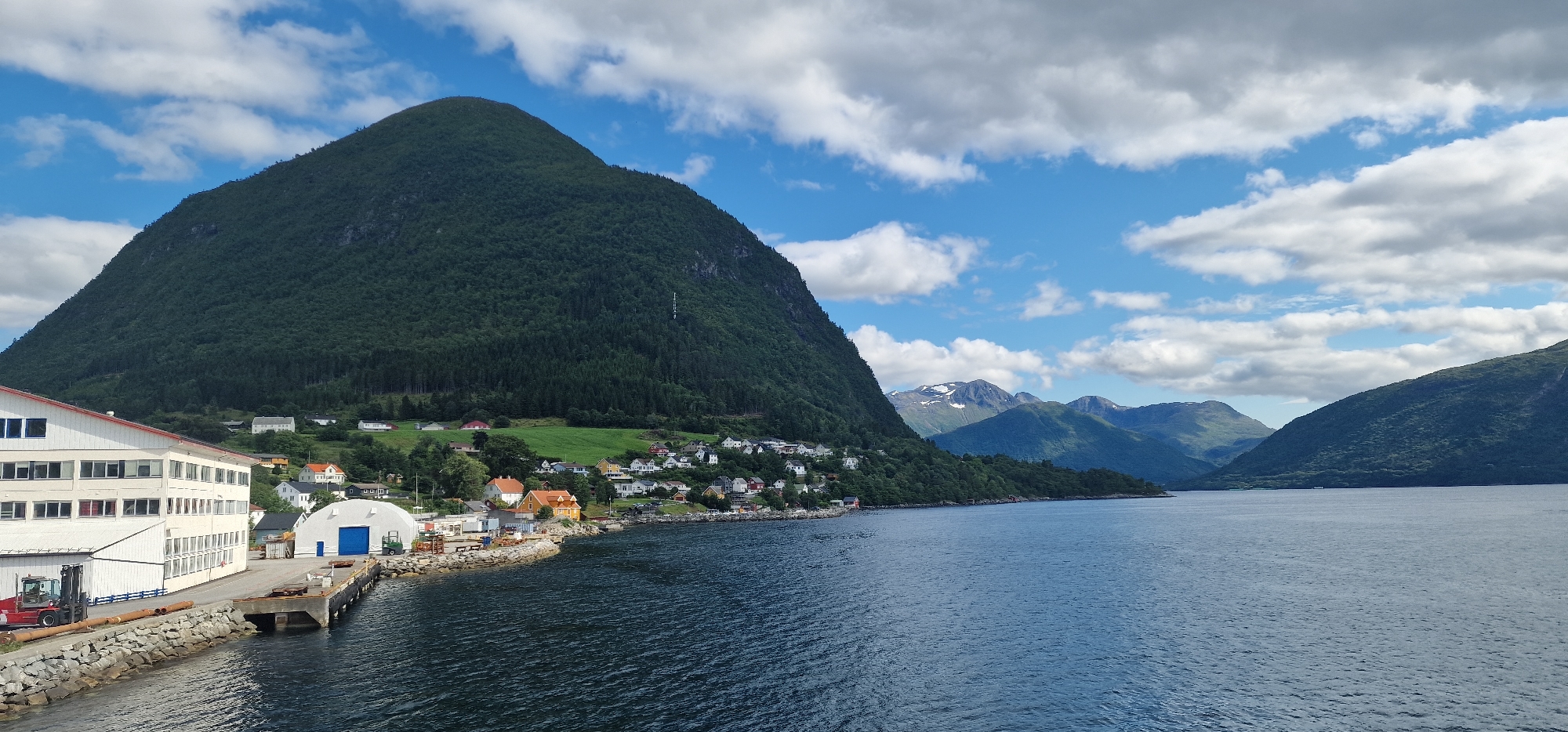 The image shows a scenic view of the city of Volda, Norway, with a focus on its harbor and surrounding landscape. In the foreground, a body of water, likely a fjord, stretches across the scene. To the left, a prominent industrial building with a white facade and multiple windows is visible, adjacent to a stone-covered embankment. A red forklift is parked near the building, and a dock extends into the water.
Beyond the industrial structures, a charming residential area is nestled on the slope of a large, verdant mountain. The houses are a mix of colors, including white, orange, and yellow, with red roofs, creating a picturesque contrast with the lush greenery. The mountain itself is densely covered with trees, with a clear cut on its upper portion that creates a dramatic visual.
In the background, a series of majestic mountains rise, some of which are capped with snow, adding to the dramatic and rugged beauty of the Norwegian landscape. The sky above is a vibrant blue, dotted with fluffy white clouds, suggesting a pleasant day. The overall impression is one of a tranquil and picturesque settlement embraced by the powerful natural beauty of the Norwegian fjords and mountains.