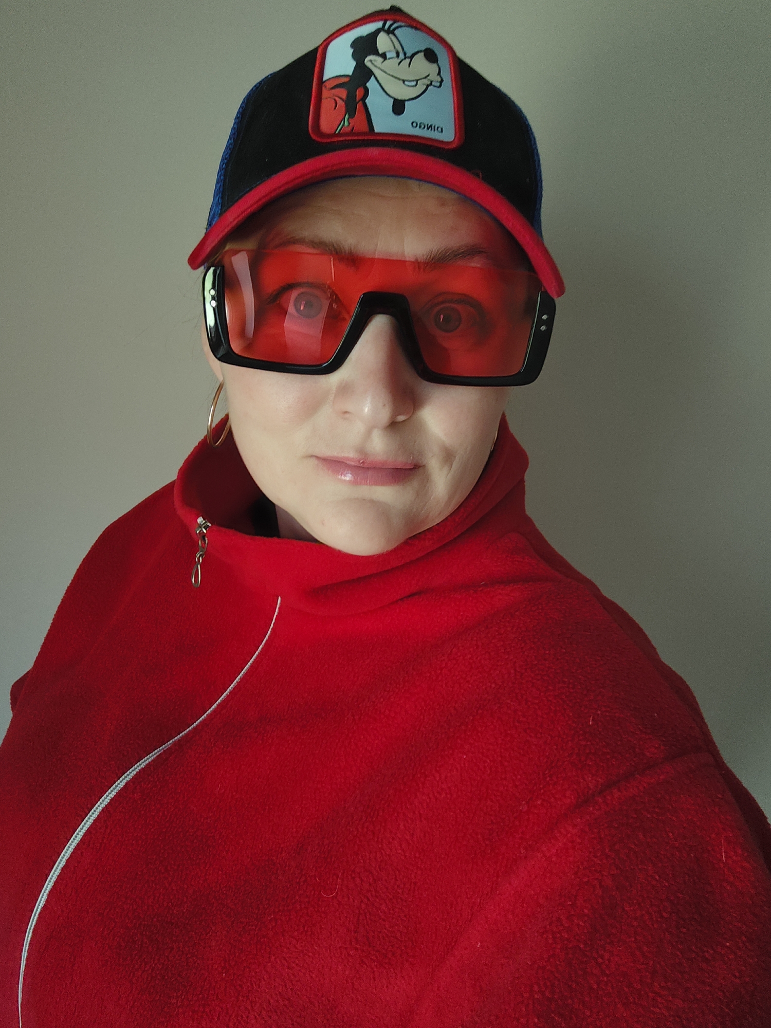 A person wearing a red fleece jacket, red and black trucker hat with a cartoon character (Goofy) patch, and large red sunglasses is standing indoors against a neutral wall. They have fair skin, light hair, and are wearing hoop earrings. The jacket is zipped partway up, revealing a dark-colored undershirt. The person is looking at the camera with a slight smile. The photo has good lighting and is in focus. There is no indication in the image that the location is in the city of Markovskii, Russia.
