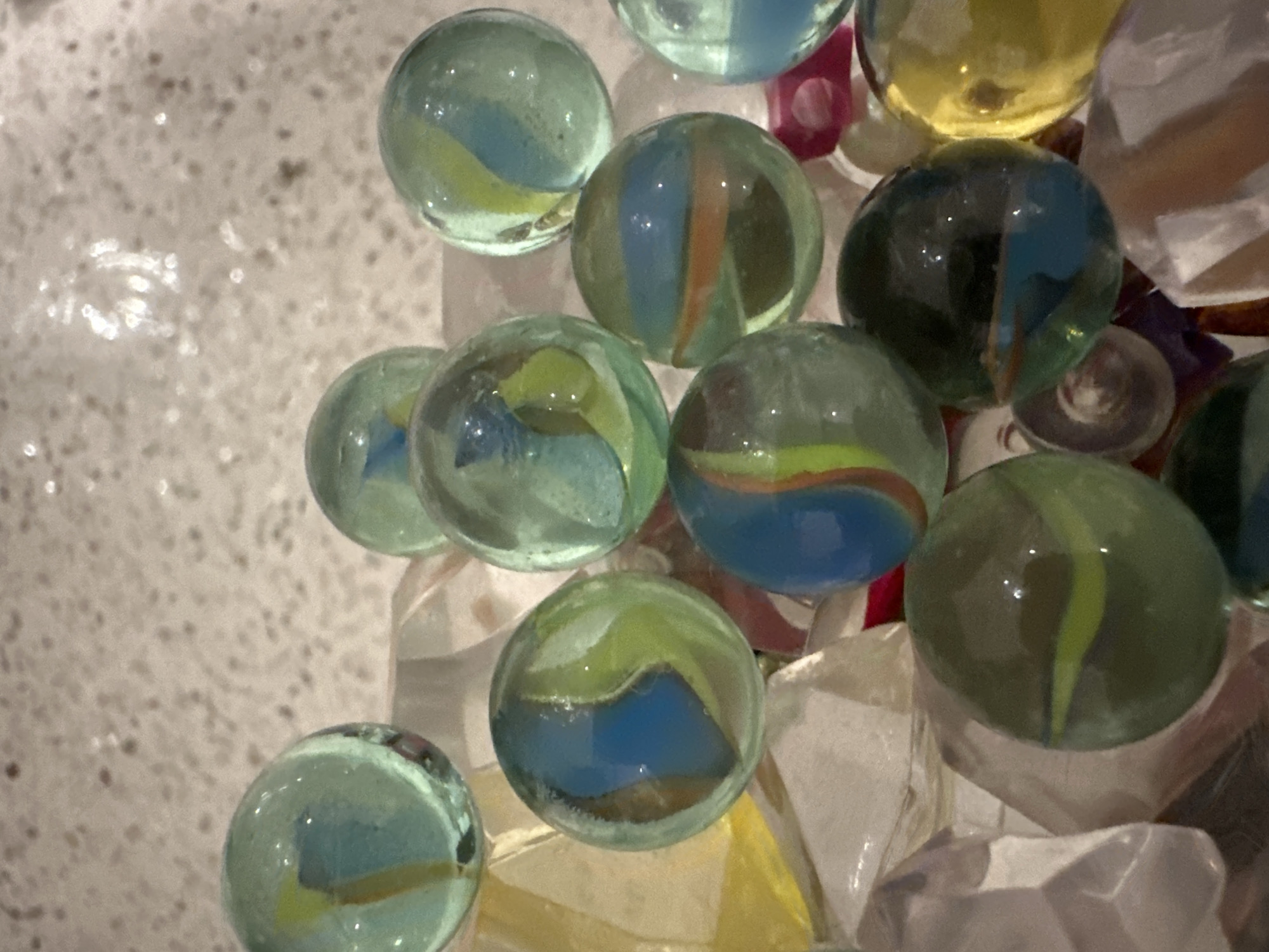 A close-up, overhead shot captures a collection of colorful marbles nestled amongst clear, faceted glass pieces. The marbles are primarily translucent shades of green, blue, and yellow, with swirling patterns of contrasting colors within. Some marbles exhibit a pale green hue with streaks of yellow and blue, while others are a deeper green with blue and orange bands. A few marbles are a vibrant yellow, and one appears to be a dark, almost black, green with blue accents. The glass pieces, which are scattered throughout the frame, have a crystalline appearance and reflect light, adding a shimmering quality to the scene. The background is a speckled, light-colored surface, possibly a countertop or table. No people or actions are present. The scene is devoid of any context regarding location within a city.