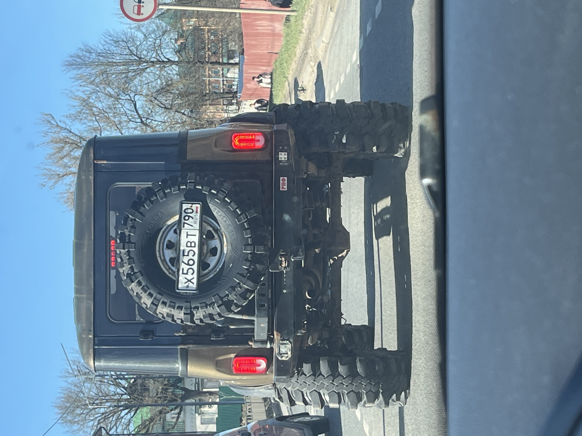 The image shows the back of a dark-colored SUV with a spare tire mounted on the rear. The license plate is visible and reads "X565BT 790 RUS". The SUV is on a road in what appears to be a city environment with buildings and trees in the background. A road sign is visible in the background. It is unclear if the location is specifically in Klin, Russia, but the license plate indicates that the vehicle is registered in Russia. There are pedestrians walking on the other side of the road.