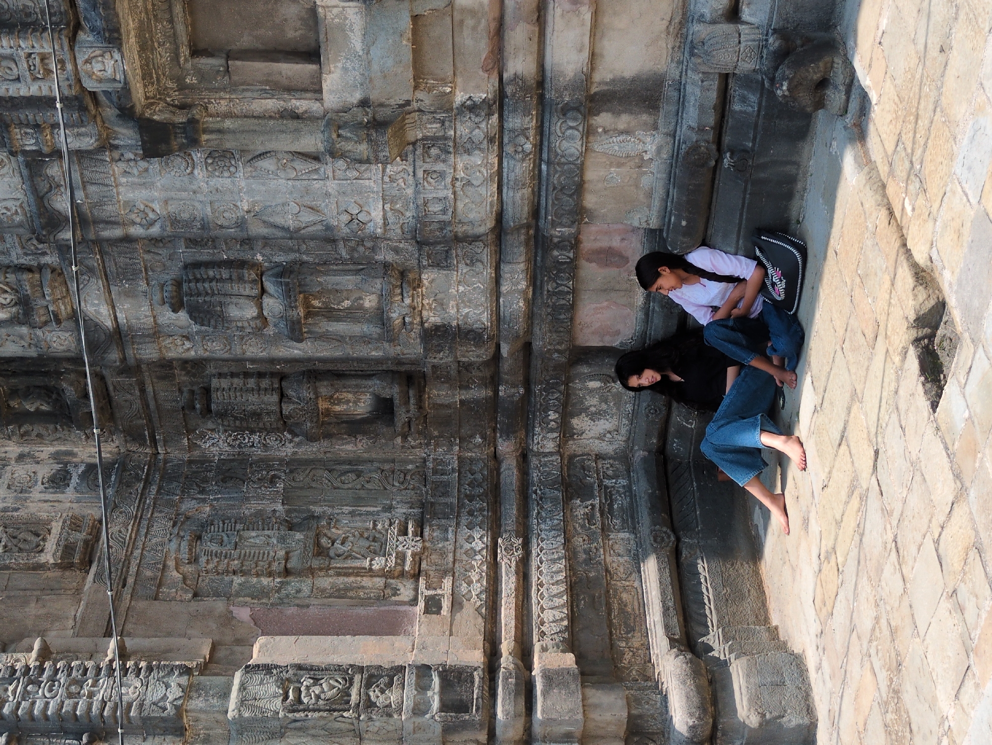 Two young women sit on a stone-paved courtyard in front of an intricately carved ancient stone temple structure. The temple wall features detailed reliefs depicting figures and decorative patterns. The woman on the left wears a black top and blue wide-leg pants, while the woman on the right wears a white long-sleeved shirt and blue jeans. A black and white patterned bag rests between them. The woman on the left is looking to her left, and the woman on the right is looking towards the viewer with a slight smile. The scene is illuminated by daylight, casting shadows on the courtyard. This is likely a location in Mandi, India, known for its ancient temples.