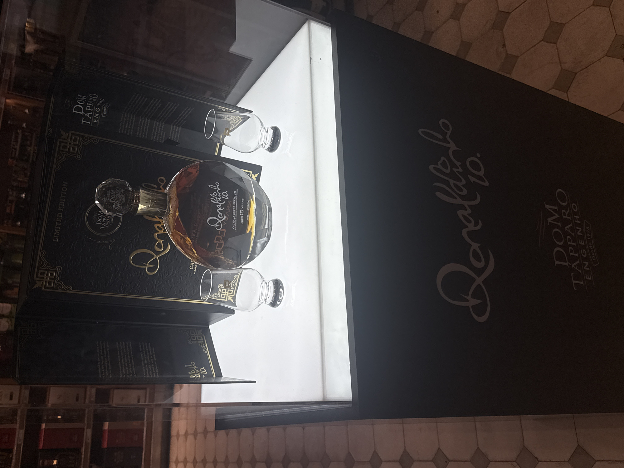 The scene depicts a product display in a retail environment in Guarulhos, Brazil. A dark-colored display stand, branded with "Ronaldinho 10." in white cursive script and "DOM TAPPARO ENGENHO Desde 1978," supports a brightly illuminated white platform. On this platform, a clear, round bottle of "Ronaldinho 10. CACHAÇA EXTRA PREMIUM AGED 10 YEARS 700 ml 40% Alc./Vol." containing an amber liquid is prominently displayed. The bottle features a faceted clear stopper. Flanking the bottle are two empty clear tasting glasses, each adorned with a gold geometric pattern near the rim. Behind the bottle, an open, black, gold-accented presentation box, labeled "LIMITED EDITION" and "DOM TAPPARO ENGENHO Desde 1978," stands upright. The interior of the box reveals partially legible text, including mentions of Ronaldinho. The background, visible through a glass partition, shows dimly lit shelves stocked with various bottles, characteristic of a liquor store or duty-free shop. The floor in the foreground features a patterned tile design with repeating hexagonal and square shapes in light and dark brown tones. No individuals are present in the image.