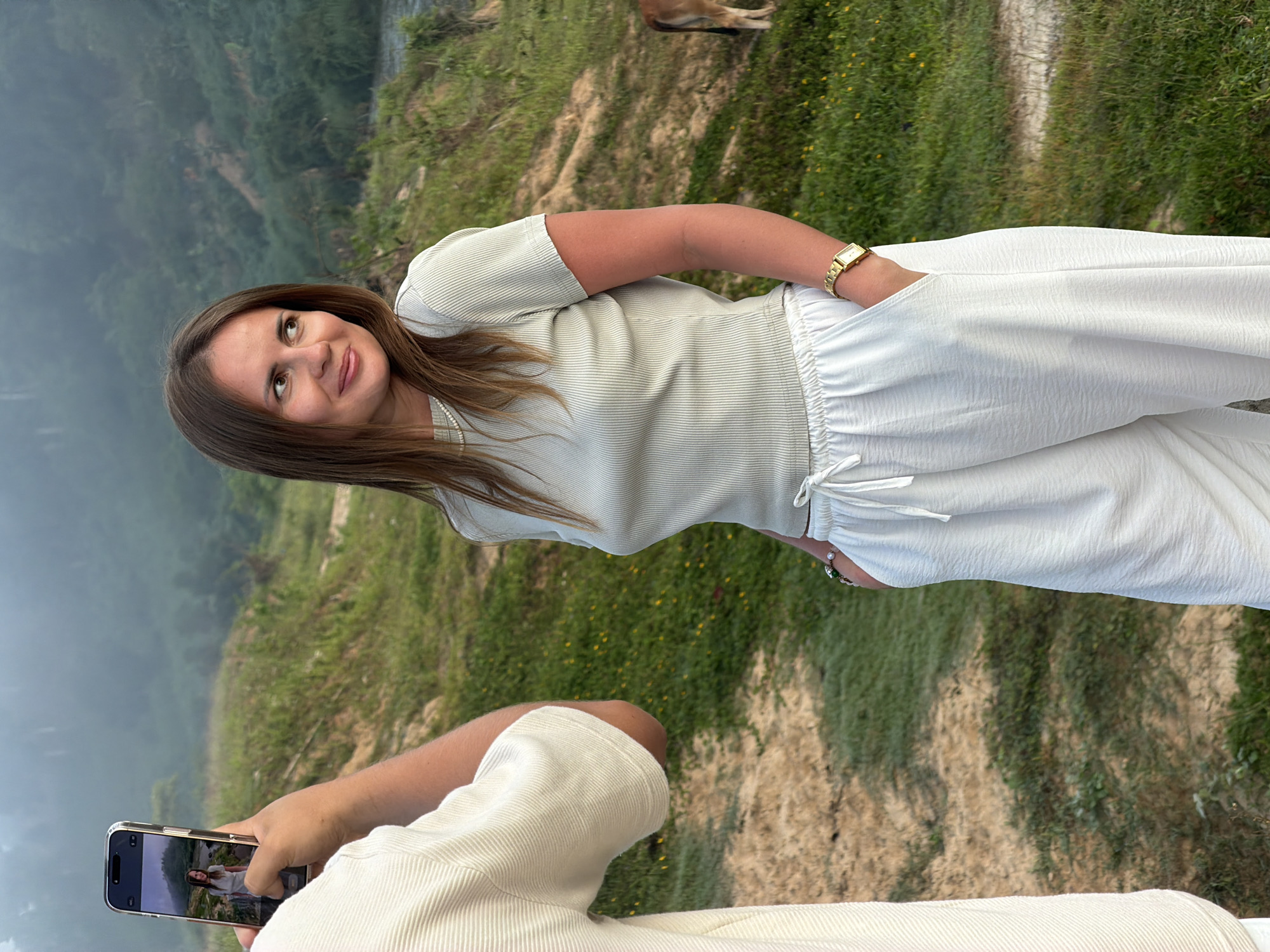 In an outdoor setting with a hazy, mountainous backdrop, a woman with long brown hair, wearing a light grey ribbed t-shirt and loose white pants, stands with her hands in her pockets. She is looking to her right, away from the camera. A person, whose arm and torso are visible from the shoulder to mid-thigh, is holding a smartphone in their right hand, taking a picture of the woman. The phone screen displays a photograph of the woman in the same pose. The ground is covered in dry grass and some scattered yellow flowers, with a path running through it. A brown cow is partially visible in the background to the right of the woman. The location is specified as Nam Khanh Vinh, Vietnam.