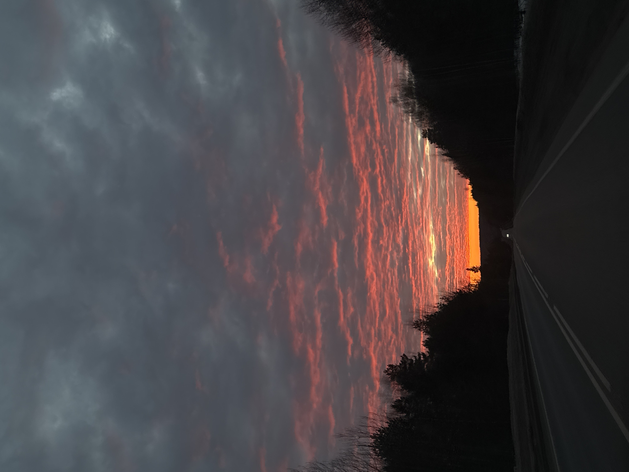 The image displays a two-lane asphalt road at dusk or dawn, looking towards the horizon in Chaikovsky, Russia. The sky features extensive dark grey altocumulus or stratocumulus clouds, exhibiting a distinct red-orange hue along the lower horizon, fading into dark grey at the zenith. The sun's intense orange glow is visible at the distant horizon line, partially obscured by terrain and trees. The road surface is dark, with a clear double solid white centerline and a single solid white shoulder line on the right, receding into the distance. Dense, dark forest, primarily composed of coniferous trees with some bare deciduous trees, lines both sides of the road, appearing in silhouette against the sky. Patches of snow are evident on the ground along the road's edges, particularly on the right. In the far distance, two faint headlights from an approaching vehicle are discernible on the road. No individuals or direct interactions are visible.