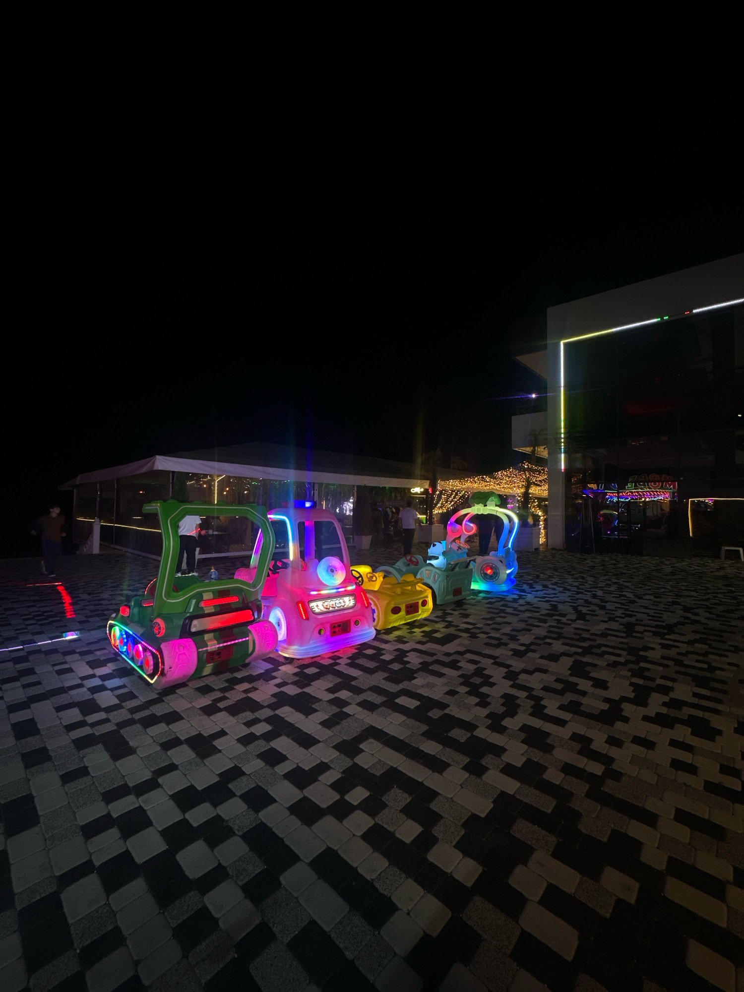 An exterior, low-angle, full shot shows a row of brightly lit, colorful children's amusement rides positioned on a patterned pavement at night. The rides are designed as vehicles, including a green tank-like vehicle in the foreground, followed by a pink car-like vehicle, and then a yellow and blue vehicle. These rides are illuminated with neon lights in various colors such as pink, blue, green, and yellow, creating a vibrant and festive atmosphere.
In the background, a white tent structure with illuminated string lights spans across the scene, suggesting an outdoor dining or event area. Beyond this, a modern building with illuminated lines of light in green, yellow, and white is visible, along with other illuminated structures and signs that are difficult to discern in detail.
A few individuals are present in the scene. One person, wearing dark clothing, is walking to the left of the frame on the patterned pavement. Another person is standing near the pink ride. Two figures are visible inside the tent structure.
The pavement is constructed from dark and light grey rectangular pavers arranged in a geometric, checkerboard-like pattern. The overall lighting of the scene is dim due to it being nighttime, with the primary illumination coming from the amusement rides and the surrounding buildings and structures.
The location is identified as Lermontovo, Russia. The scene appears to be a recreational area, possibly a park or an entertainment venue near a beach or waterfront, given the presence of the tent structure often seen in such locations. The specific time is night.
