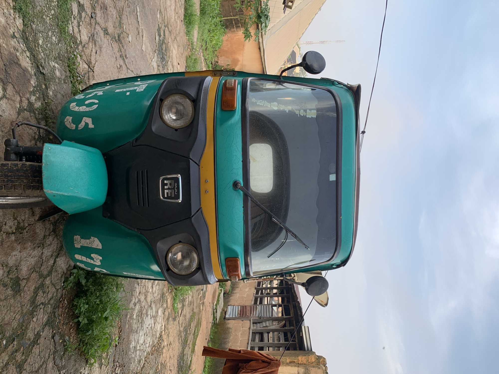 A turquoise and yellow three-wheeled auto-rickshaw is parked on a cracked asphalt surface in a residential area of Jos, Nigeria. The auto-rickshaw faces the camera, with its windshield reflecting the cloudy sky. On the left side of the auto-rickshaw, the number "17595" is stenciled in white. On the right side, the number "17" is partially visible, also in white. The background shows a brick building with corrugated metal roofing, and an orange jacket is hanging on a clothesline. Green vegetation and a small tree are visible to the left of the auto-rickshaw. The sky is overcast with a few patches of blue.