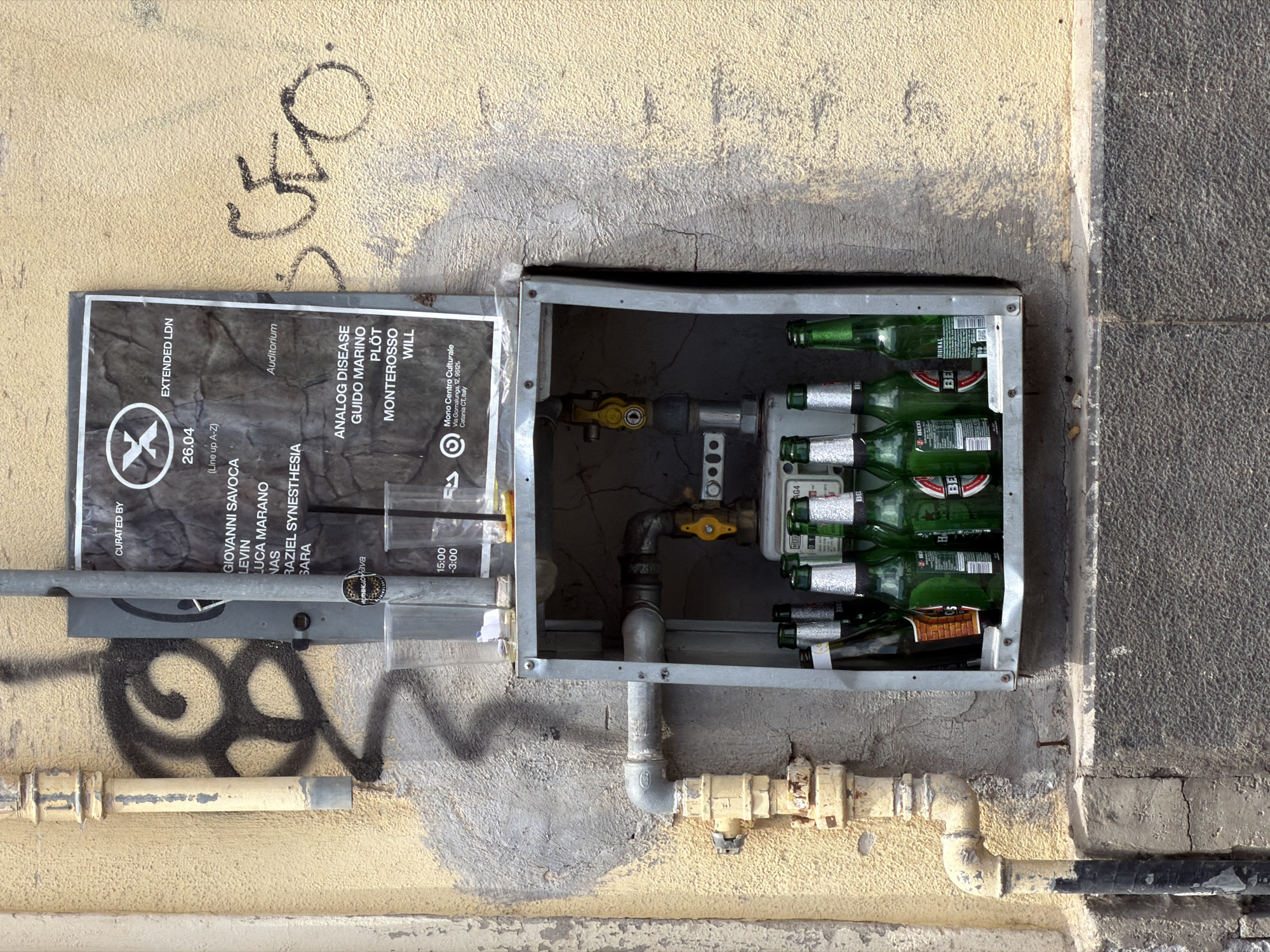 This image depicts a utility box mounted on a weathered exterior wall in Catania, Italy. The wall has a multi-textured surface with areas of stucco, concrete, and exposed aggregate. Patches of faded yellow paint are evident, along with black graffiti. A metal utility box is partially open, revealing a complex arrangement of pipes, valves, and a gas meter. Several green beer bottles have been crammed into the box alongside the mechanical components. Affixed to the box's door is a black and white poster for an event titled "Extended LDN" at the Mono Centro Culturale (Via Gornalunga, 12, 95126 Catania CT, Italy) on April 26th, featuring a lineup of artists. A pipe runs horizontally along the wall beneath the utility box.