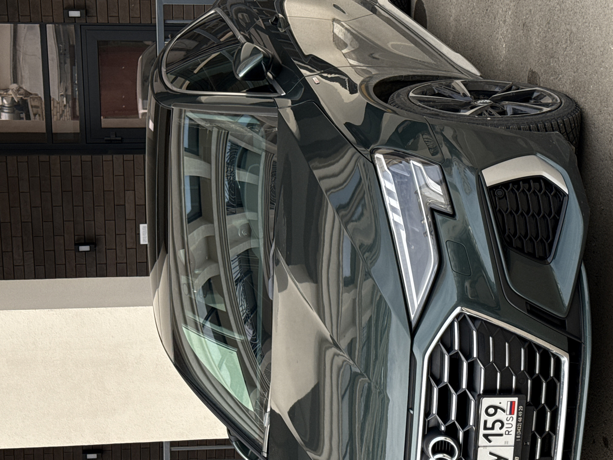 The image shows the front of a dark green Audi car parked on an asphalt surface. The car has an Audi logo and a Russian license plate with the numbers "Y 159". In the background, there is a building with windows and a brick facade. The photograph appears to have been taken in Perm, Russia, as indicated by the Russian license plate.