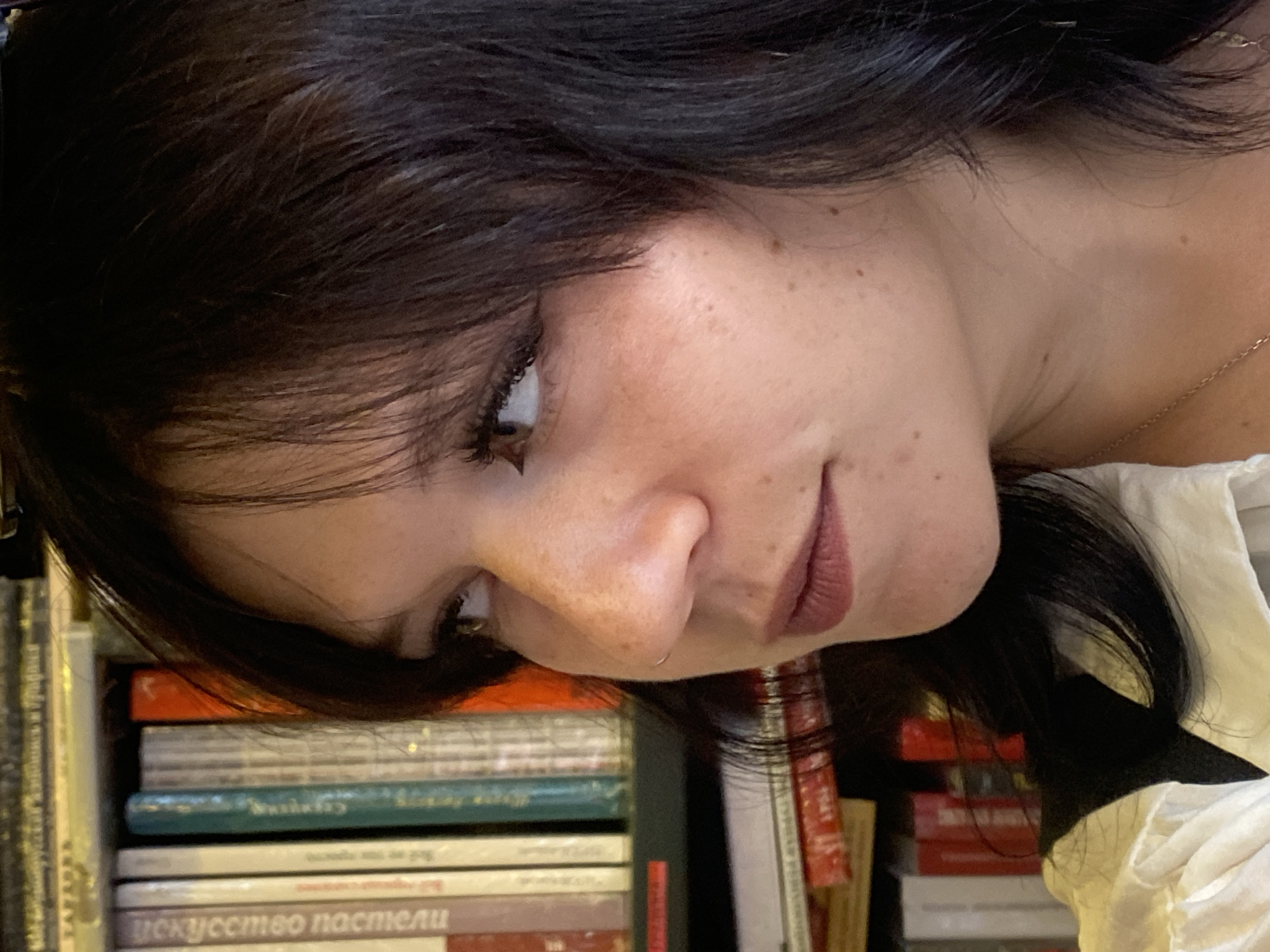 A close-up, profile shot of a young woman with dark hair and fair skin with freckles is featured. She is looking to her left. Her makeup includes winged eyeliner and a muted lipstick. She is wearing a white collared shirt and a dark vest or strap. A thin necklace with a pendant is visible. Behind her, bookshelves filled with books are visible. The image appears to have been taken indoors, likely in a home or library setting, given the presence of bookshelves. The specific location in Saint Petersburg, Russia, is not discernible from the visual information provided in the image.