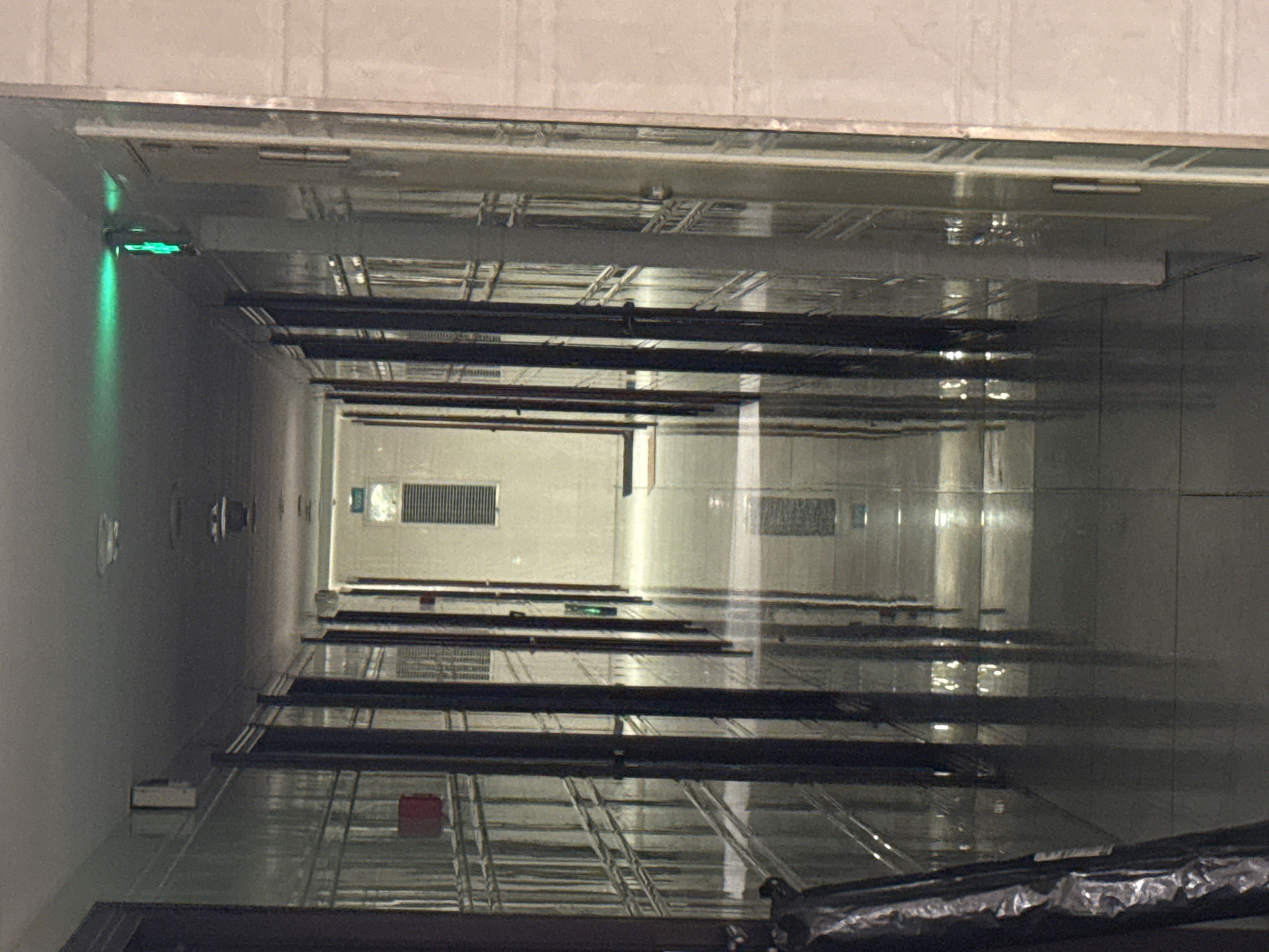 The image shows an upward view inside a building's elevator shaft. The shaft walls appear to be made of a reflective, possibly metallic, material. There are multiple floors visible, separated by structural supports and elevator doors. A green light is visible at the top of the shaft on the left side. The location is Shapingba District, China.