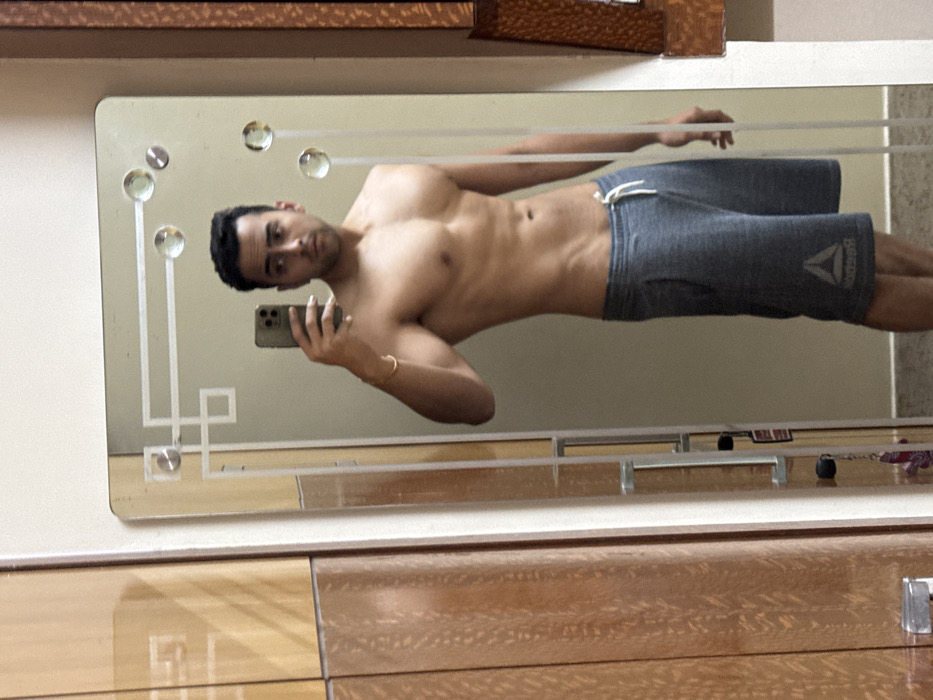 A man in gray Reebok shorts takes a selfie in a mirror. He's shirtless, revealing a toned physique. The mirror has a vintage design with geometric patterns and spherical embellishments. The background includes a wooden dresser or cabinet. The man holds his phone with his right hand.