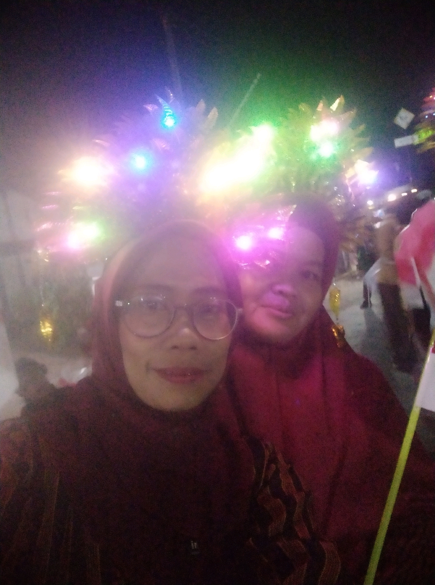 The image depicts a nighttime outdoor scene likely at a festival or celebration in Karanggayam, Indonesia, characterized by numerous bright, colorful lights that illuminate the background. In the foreground, two individuals are visible. The person on the left, wearing glasses and a headscarf, is positioned closer to the camera, their face is well-lit. To their right, another person, also wearing a head covering, is partially visible. The clothing of both individuals appears to be dark, possibly red or maroon. The overall atmosphere is festive and somewhat blurry due to the low light conditions and potential movement during the photograph.