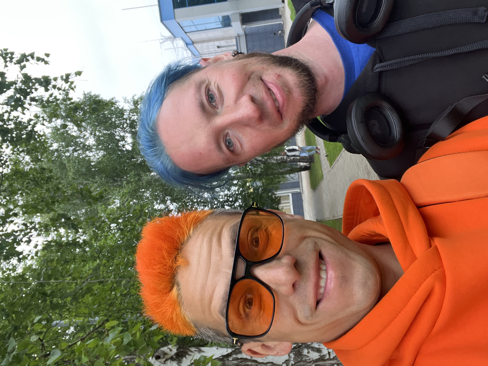 Two men are posing for a selfie outdoors. One man has blue hair and a beard, and wears a blue shirt under a black hoodie with headphones around his neck. The other man has orange hair and orange-tinted sunglasses, wearing an orange hoodie. They are both smiling. In the background, there are trees, a sidewalk, and a modern building with glass windows. The location appears to be in the city of Chaikovsky, Russia.