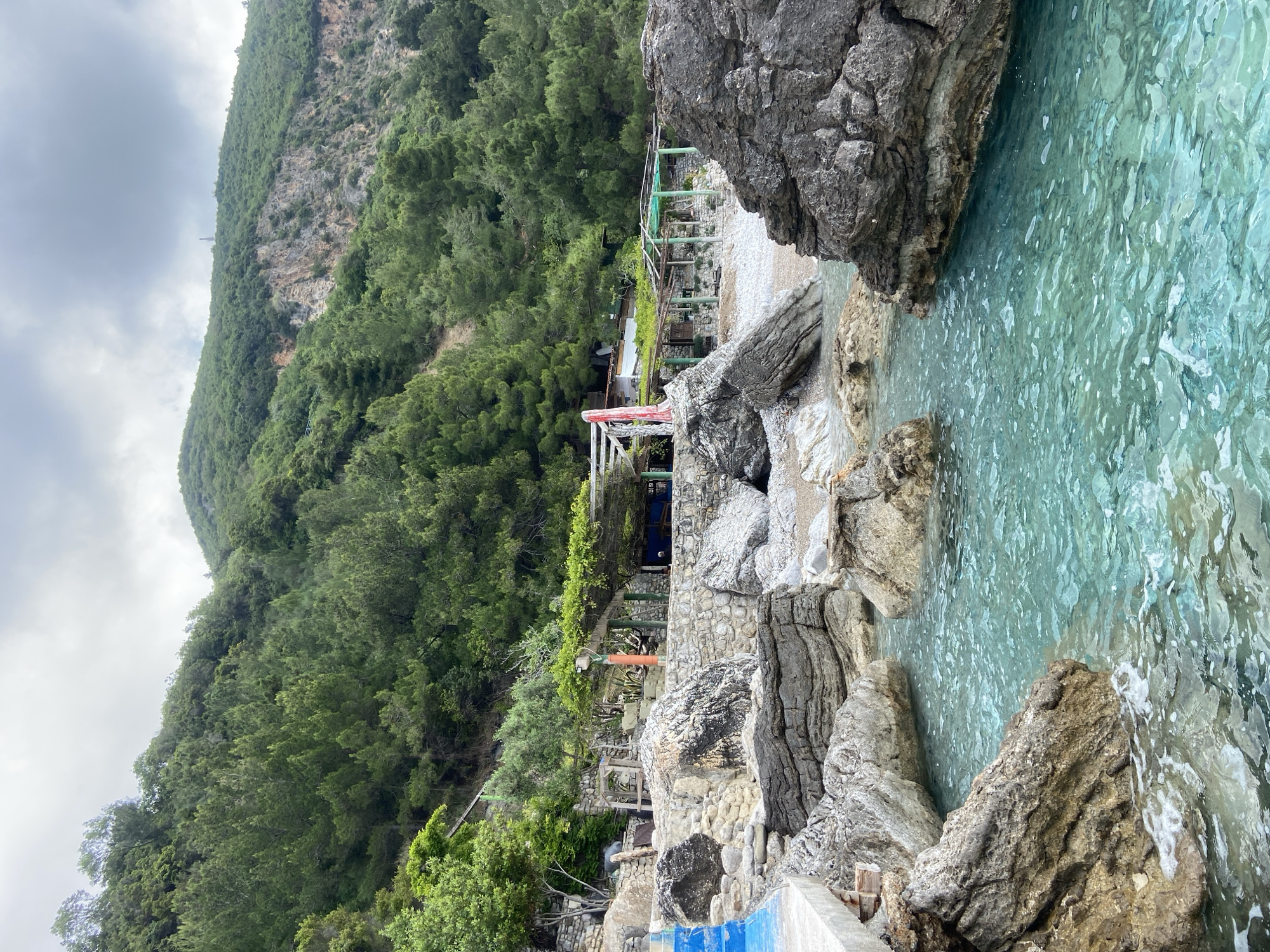 The image captures a coastal scene in Prijevor, Montenegro. Clear turquoise water fills a small cove bordered by large, rough-textured gray rocks. Vegetation, including trees and shrubs, covers the hillside behind the beach. A stone structure with green supports and a wooden trellis is visible along the shoreline. A steep, forested mountain rises in the background under a cloudy sky.