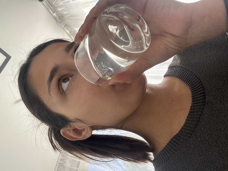 The scene, located in Budva, Montenegro, depicts an indoor environment. A female individual, shown from the mid-chest upward, is visible. She has dark hair pulled back into a ponytail. Her right eye is directed upwards and to the right. She wears a dark gray knitted sweater. Her right hand holds a clear, textured glass tumbler filled with transparent liquid, likely water, to her mouth, indicating the act of drinking. The blurred background features a bright window on the left, partially obscured by sheer white curtains, and a dark, angular object against a white ceiling.
