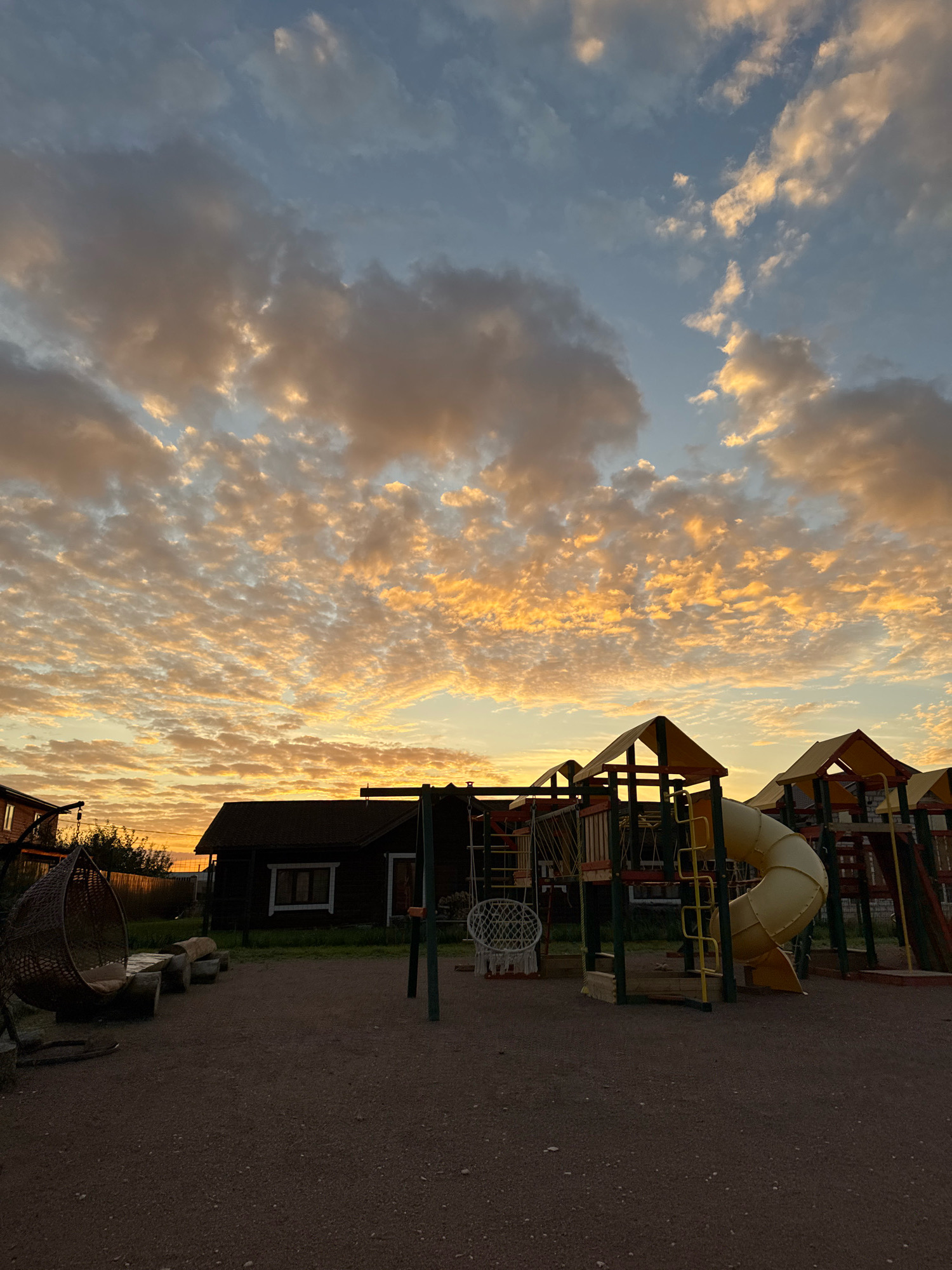 The image depicts an outdoor playground and a dark wooden house under a dramatic sunset sky filled with mottled orange and yellow clouds. The playground features a yellow tube slide, climbing structures, and a basketball hoop. To the left of the house, there is a large, dark wicker hanging chair. The ground is covered in gravel. The scene is set in the city of Kempelevo, Russia. No people are present in the image, and there are no observable actions or interactions taking place. The environment appears to be a rural or suburban setting.