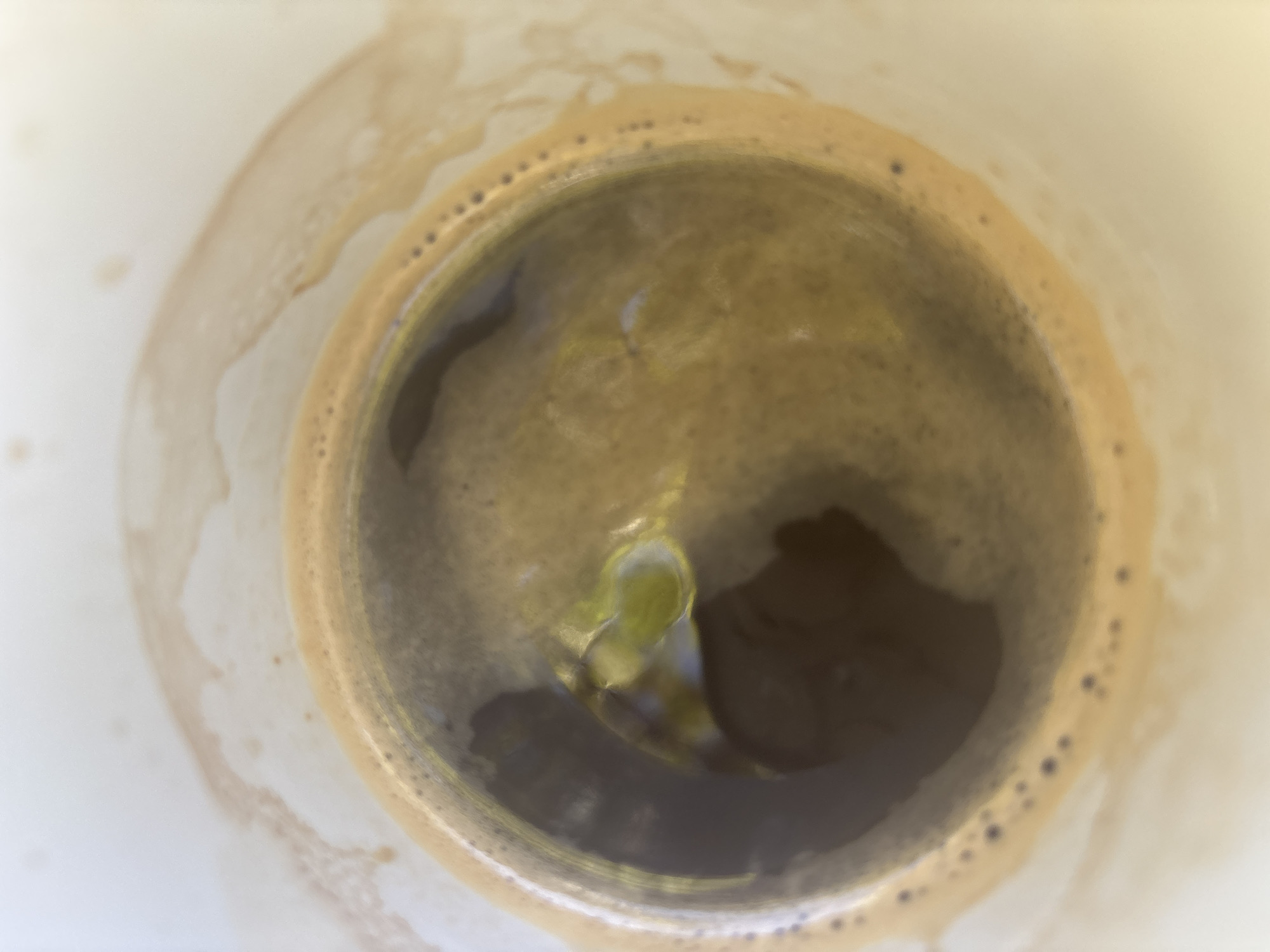 The image is a close-up, overhead view of a cup of coffee. The coffee has a frothy, tan-colored crema around the edges and a darker liquid below. There are some light brown stains on the white surface surrounding the cup. The scene is generic and does not contain any elements that would allow for identification of a specific location in Trudovets, Bulgaria, or any other city. There are no people, animals, or distinct landmarks visible. The focus is solely on the coffee and the cup.
