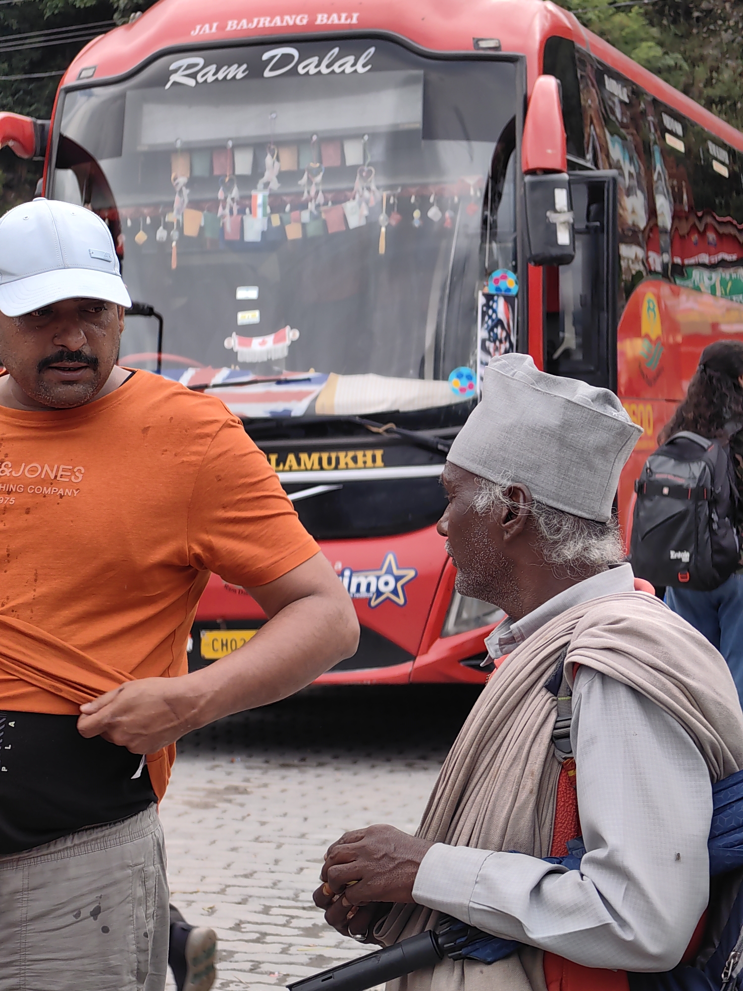The image shows a street scene in Siyal, India. Two men are in the foreground, standing near a red bus. The man on the left is wearing an orange t-shirt and a white baseball cap, and is pulling at the hem of his shirt. The man on the right is wearing a grey cap and a light-colored shirt, with a tan shawl draped around his shoulders. He appears to be sitting or leaning, possibly in a wheelchair, and is holding something in his hands. The bus behind them has writing on it that includes "JAI BAJRANG BALI" and "Ram Dalal". Small decorative flags are hanging from the bus's interior. The ground appears to be paved with cobblestones or similar material, and some trees are visible in the background. A backpack is visible on another person further back in the image.