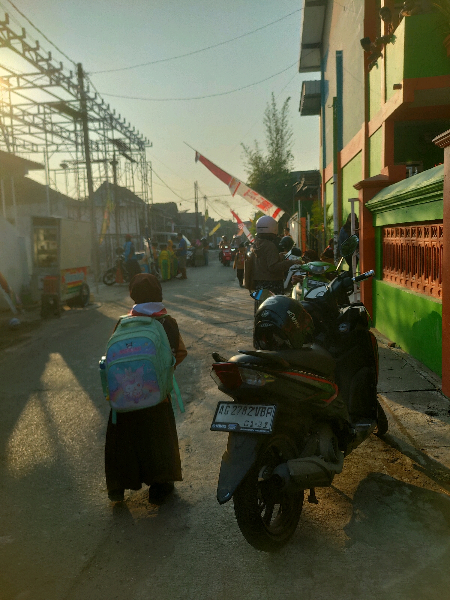 In the city of Nganjuk, Indonesia, an image captures a street scene during what appears to be late afternoon or early morning, indicated by the warm, low-angle sunlight casting long shadows. The foreground is dominated by the rear view of a child wearing a dark headscarf and a brown school uniform, carrying a light blue backpack adorned with cartoon characters. The child is standing on a paved street, facing away from the camera and towards a row of buildings and parked motorcycles.
To the right of the child, a dark-colored motorcycle is parked, its license plate reading "AG 2782 VBR". A black helmet rests on its seat. Further down the street, other motorcycles are visible, along with several people in the background. One person, wearing a helmet and a light brown jacket, is partially visible on a motorcycle. Another individual in the background appears to be wearing a green and yellow outfit.
The buildings lining the street vary in color and architectural style. On the right, a building with green and peach-colored walls features decorative window grilles. On the left, a structure with scaffolding suggests ongoing construction or renovation. Red and white flags, likely Indonesian national flags, are displayed along the street, hinting at a national holiday or celebration. The sky is clear and bright, with some power lines and poles visible. The overall atmosphere is one of everyday life in a bustling urban environment, with a touch of festive decoration.