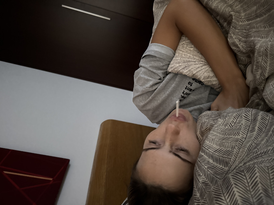 A person, appearing female, is observed lying on their left side in a bed within an indoor room in Warsaw, Poland. The individual has dark hair and closed eyes, with a white, cylindrical object protruding from their mouth. They are dressed in a grey short-sleeved t-shirt with partial text visible on the chest, including "re." and "ant want to come". The person is partially covered by a duvet exhibiting a repeating geometric pattern in shades of grey. A light-colored wooden headboard is visible behind the individual. The background features a plain white wall to the left, adorned with a reddish-brown abstract artwork incorporating golden linear elements. To the right, a dark brown wardrobe or cabinet with a single vertical silver handle is present. The room is dimly lit.