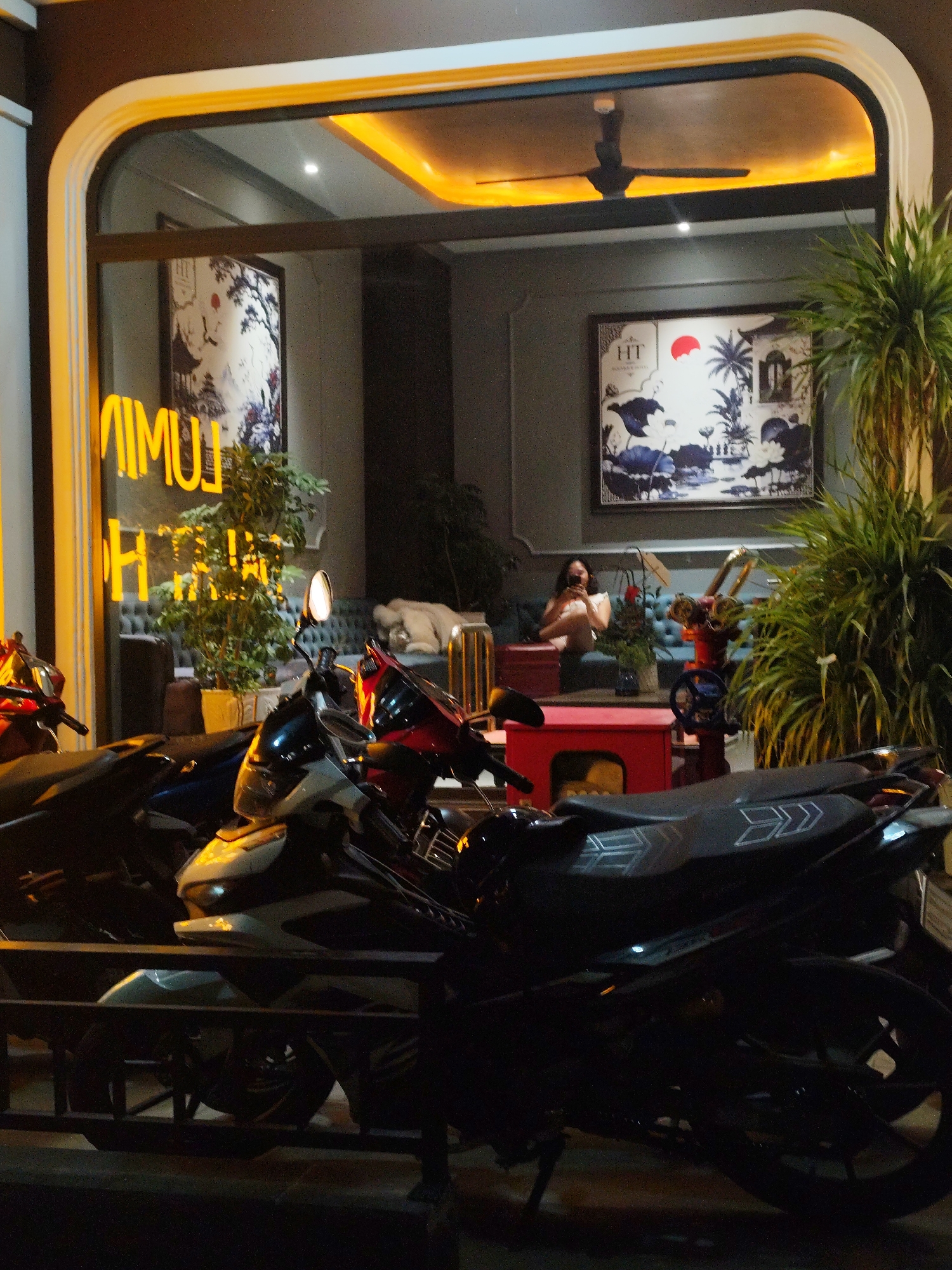 The image depicts a nighttime street scene in front of a brightly lit establishment, likely a cafe or boutique, located in Da Lat, Vietnam. The establishment's exterior features a large, curved window with illuminated yellow lettering that reads "LUMINA". Inside, the interior is dimly lit, with a ceiling fan visible and two framed artworks on the wall. One artwork displays a traditional landscape with a red sun and a bird. Another artwork is partially visible and appears to be a mural or painting.
In the foreground, several motorcycles are parked outside the establishment, obscuring a portion of the lower half of the image. The motorcycles appear to be common street models, with one prominently featuring a black seat with white arrow-like decals. A metal railing or fence runs horizontally across the lower portion of the image, separating the motorcycles from the viewer's perspective.
Inside the establishment, a woman is seated on a plush blue sofa, holding a phone and looking towards it. A red side table is positioned in front of the sofa. To the right, a large potted plant with lush green foliage adds a touch of nature to the scene. Further inside, a beige-colored object resembling a large cushion or blanket is draped over what appears to be another sofa or seating area.
The overall impression is that of a contemporary urban setting at night, with a mix of modern design and traditional artistic elements within the establishment. The presence of motorcycles suggests a public space accessible by road, common in Vietnamese cities. The yellow neon signage indicates a business aiming to attract attention in the evening.