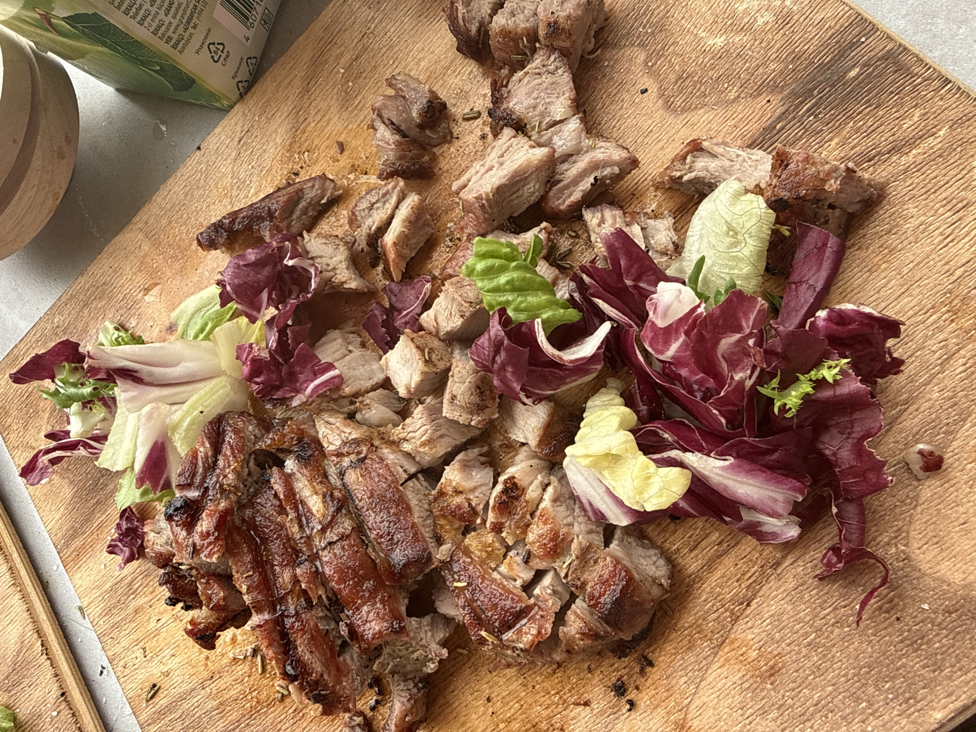 The image shows a wooden cutting board with sliced and chopped grilled meat arranged amongst a mix of red and white radicchio lettuce and herbs. The meat is the primary focus, appearing to be the remnants of a meal. Some of the meat is cut into thick strips, while the rest is roughly cubed. The radicchio appears to be a garnish. The upper left of the frame includes a glimpse of what appears to be a carton. The overall image gives a close-up, food-oriented perspective with a natural, warm lighting. There is no evidence to suggest that this photo was taken in Chaikovsky, Russia.