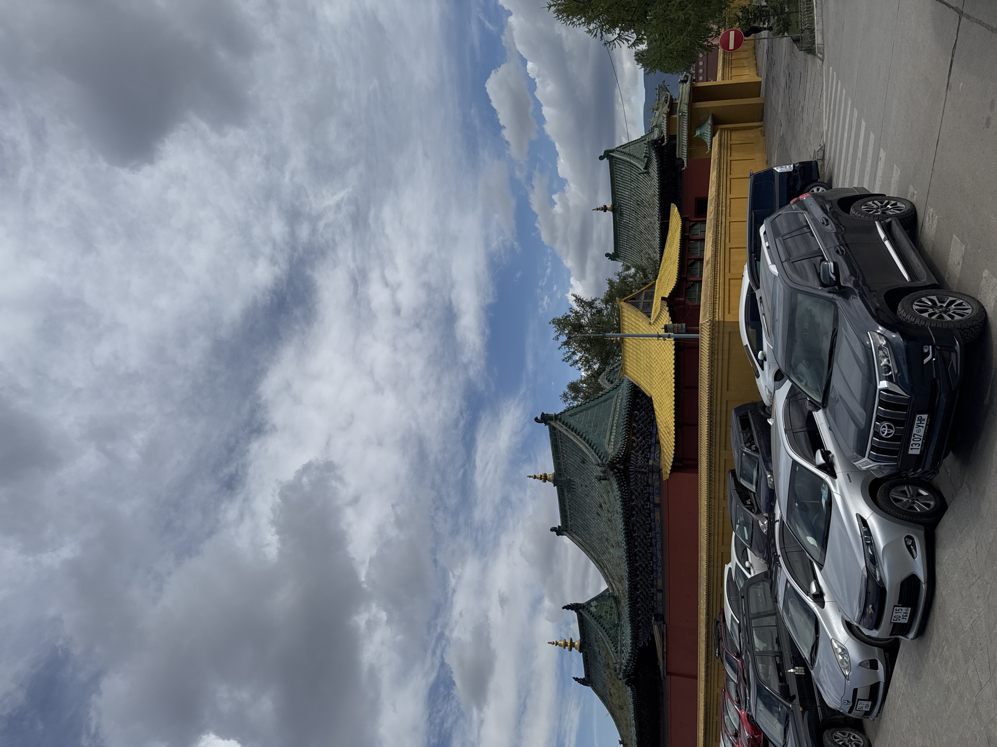 The image captures a street scene in Ulaanbaatar, Mongolia. A row of parked cars lines the right side of the frame, facing the camera. To the left of the cars is a yellow and red building with a multi-tiered roof structure, characteristic of Buddhist architecture. The sky above is partly cloudy with both white and dark clouds. The architecture and parked cars suggest a modern urban environment juxtaposed with traditional cultural elements. A zebra crossing is visible. A road sign is at the upper right of the image.