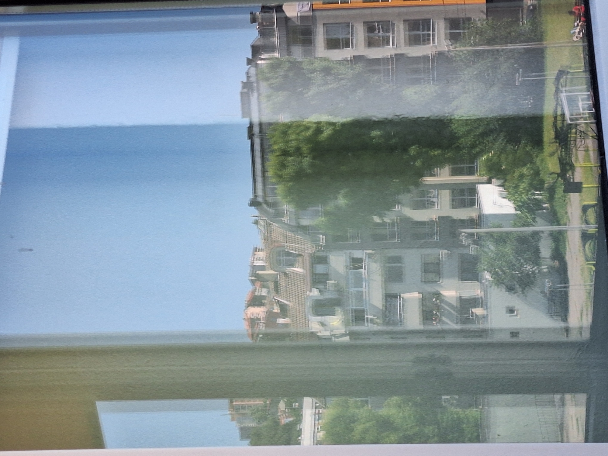 The image shows a view through a window of buildings and trees in the city of Varna, Bulgaria. The buildings are multi-story residential structures with tiled roofs and visible balconies. There are also green trees and some vegetation visible in front of and between the buildings. The sky is clear and blue. The window itself has some reflections, making the view slightly hazy and distorted in places. The scene appears to be a daytime view of a city neighborhood.