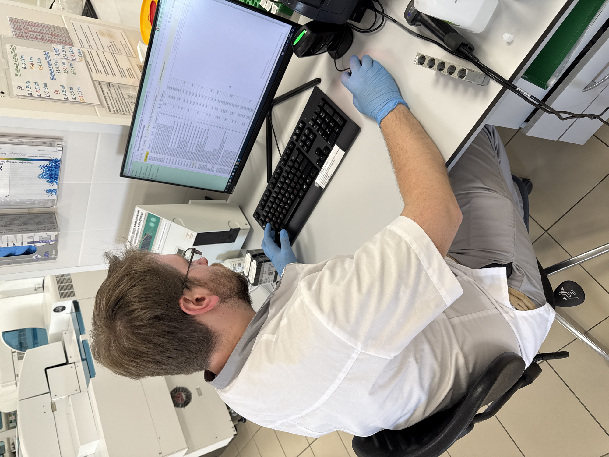 An overhead shot captures a person, likely a lab technician or scientist, working at a desk in what appears to be a laboratory or medical facility in Moscow, Russia. The individual, wearing a white lab coat and blue protective gloves, is seated in a black office chair, focused on a computer monitor displaying a spreadsheet or database with numerous rows of data. Their right hand, gloved, rests on a computer mouse, while their left hand is near the keyboard.
The workspace is equipped with a black computer keyboard and mouse, and a barcode scanner. To the person's left, a large, white laboratory instrument with blue accents is visible. Various lab supplies and equipment are scattered around the desk, including bottles, a white power strip, and what looks like a reagent holder. The floor is made of light-colored tiles, and the walls are white.
Notable visible text includes Cyrillic labels on papers taped to the wall, listing "Желчные к-ты (Ваda)" (Bile acids (Ваda)), "Zn", "Cu", "Жирные к-ты (UnBa)" (Fatty acids (UnBa)), and "AMG", followed by various reagent specifications like "R1-A 25 ml", "R2-B 4 ml", etc. A box with "NITRILE" visible suggests gloves. Other text includes "000 пљу брег" and a phone number "5-800-100-85-06". The overall scene conveys a professional, busy environment dedicated to analysis or data processing.