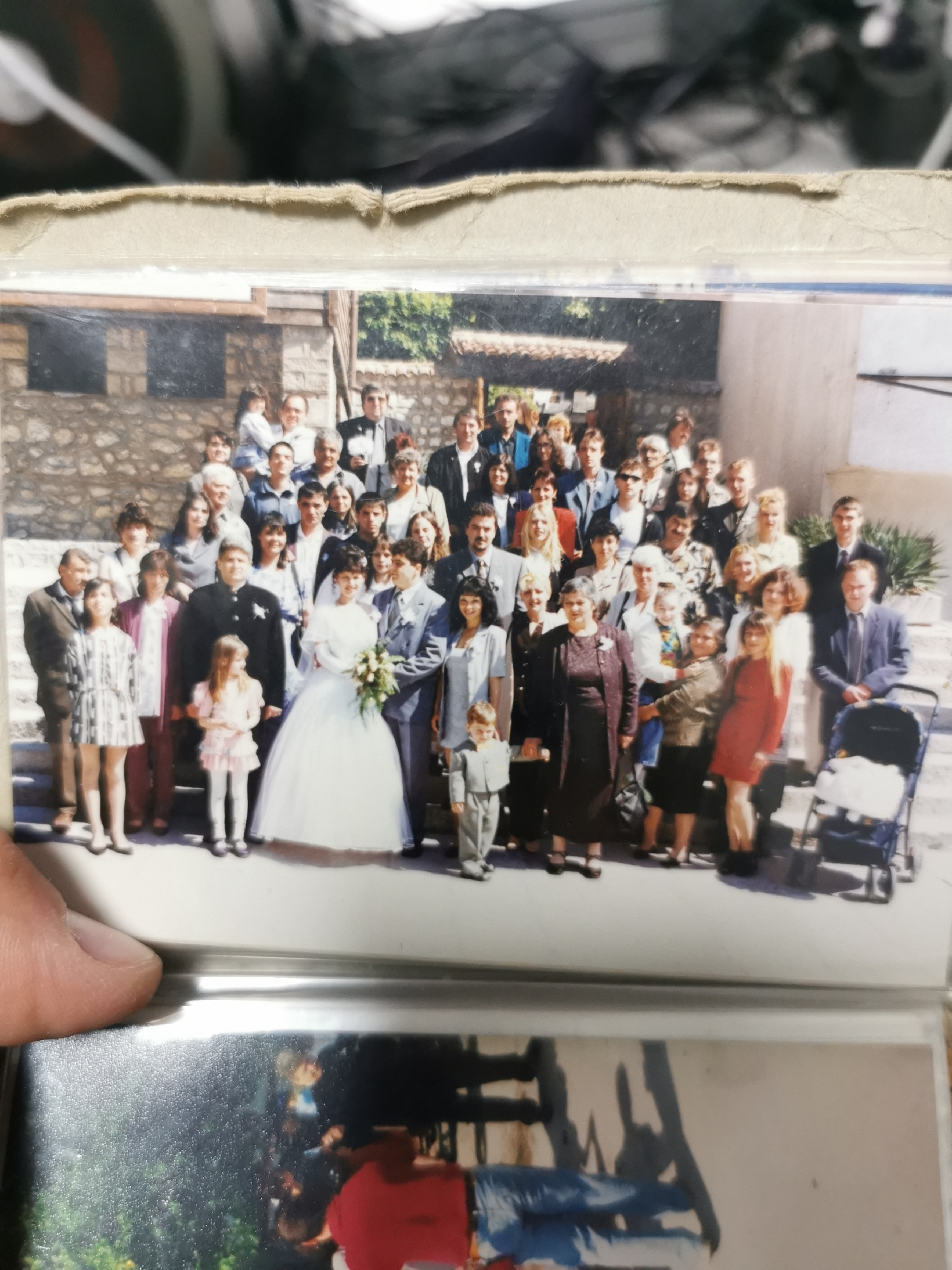 The image shows a wedding group photo taken outdoors in what appears to be Varna, Bulgaria. The group is posed on steps in front of a stone building. At the front of the group are the bride in a white gown and the groom in a suit. To their right stands a young boy in a suit. Other adults and children, presumably family and friends, are standing around them, arranged on different steps to create depth. The building in the background appears to have a stone facade and simple architecture. A baby stroller is visible near the right edge of the photo. The photo itself appears to be a snapshot from an older photo album.
