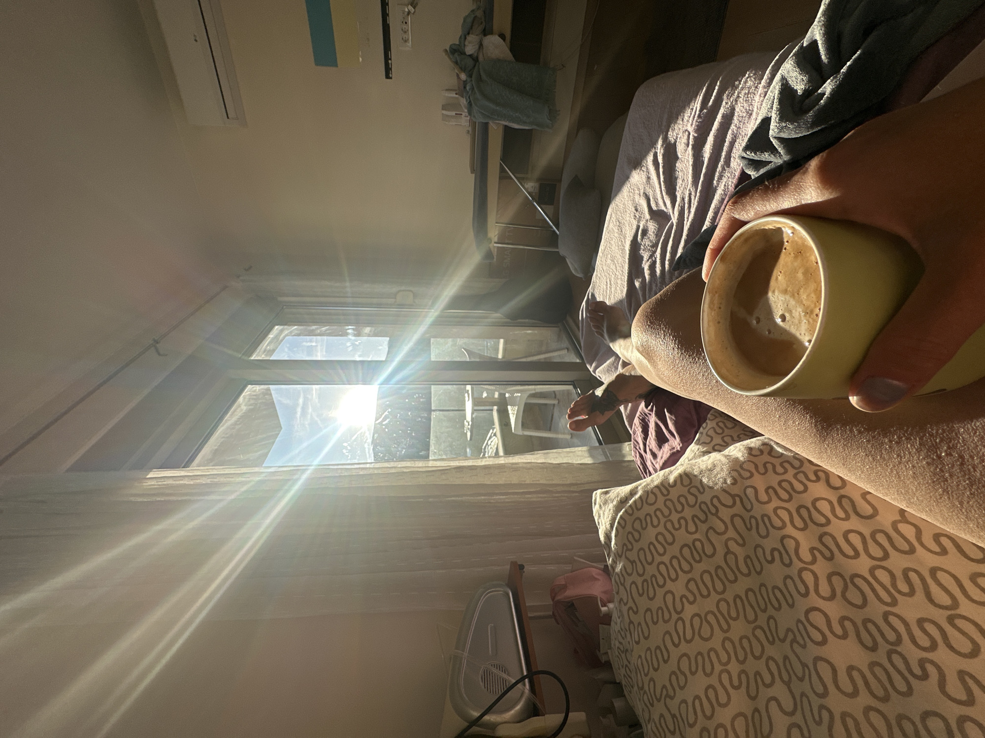 An indoor, slightly tilted shot from a low perspective, possibly from a bed. The subject is holding a mug of coffee. The view overlooks an exterior scene of Budva, Montenegro, filled with daylight. Visible are architectural elements and a balcony or terrace. The pillow with brown wave patterns in the foreground and the sunlit bedroom suggest a relaxed morning setting. The image also captures a humidifier or air purifier and parts of furniture. A glimpse of the person's leg and foot is also noticeable.