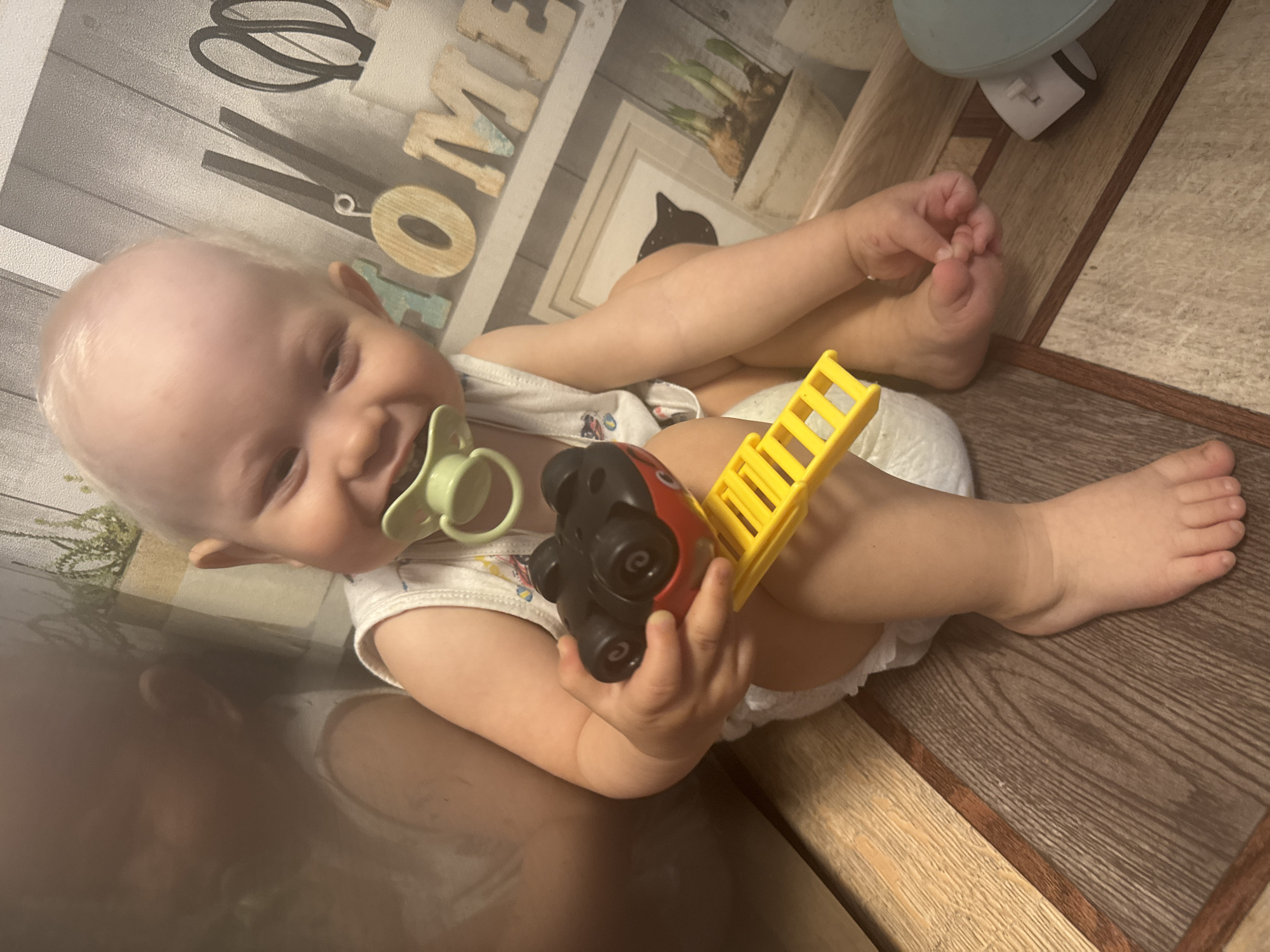 The image shows a young child, appearing to be a toddler, sitting on a wooden floor. The child has blonde hair and is wearing a white tank top and a diaper. A green pacifier is in the child's mouth, and they are holding a red and black toy with a yellow ladder. The background features a wall with a decorative wallpaper that includes the word "HOME" in wooden letters, along with images of plants and scissors. There are also potted plants and framed pictures on a shelf. To the left of the child, a reflective surface shows a distorted image of what appears to be part of a closet or a piece of furniture. The lighting suggests an indoor setting, likely a home environment. The image does not contain any information that can be used to determine its location in the city of Chaikovsky, Russia.